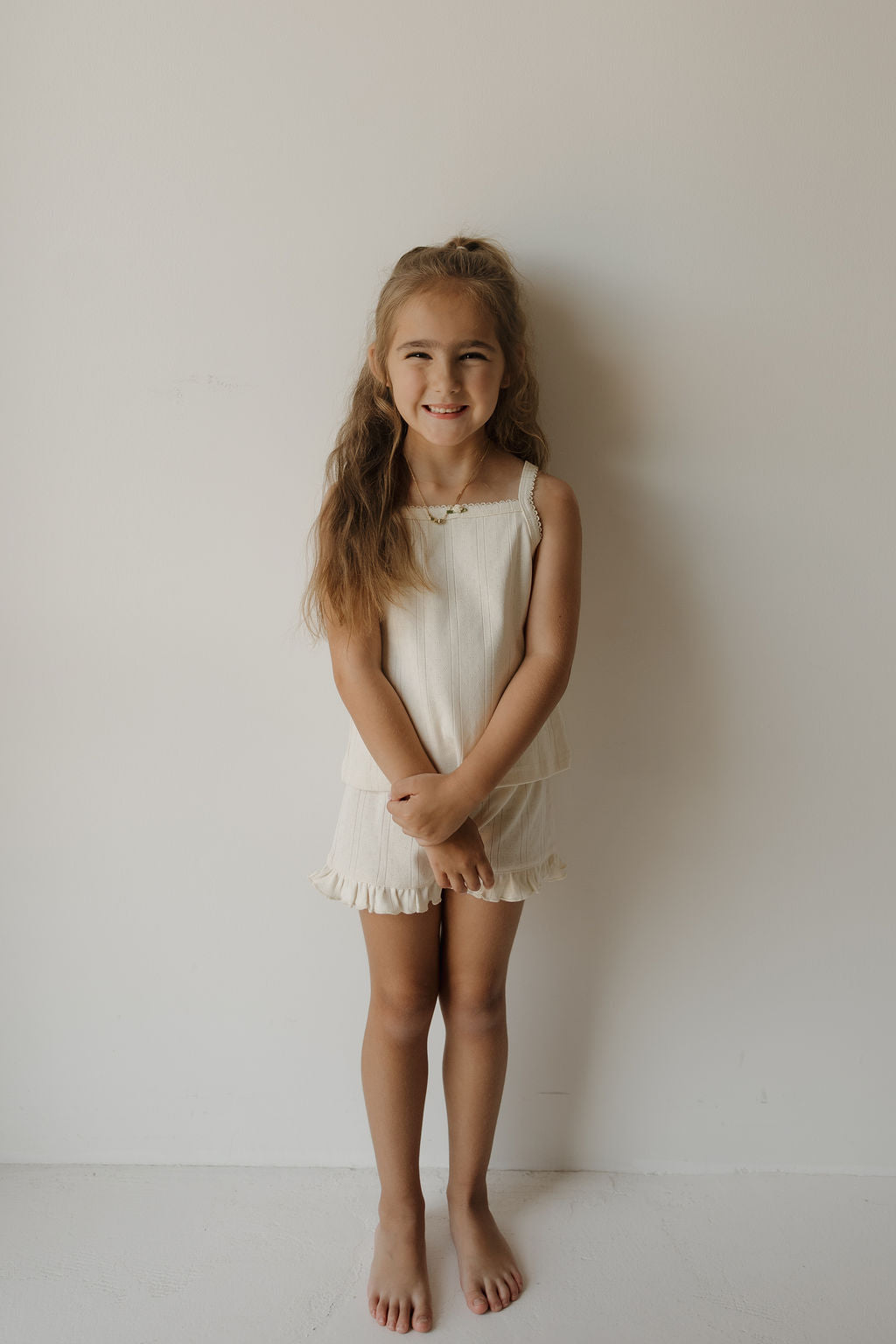 A young girl with long brown hair stands barefoot against a white wall, smiling and wearing the forever french baby Girl’s Ruffle Short Set | Swan—a sleeveless cream-colored top and matching ruffle shorts crafted from soft pointelle fabric—making it a charming summer outfit perfect for warm days.