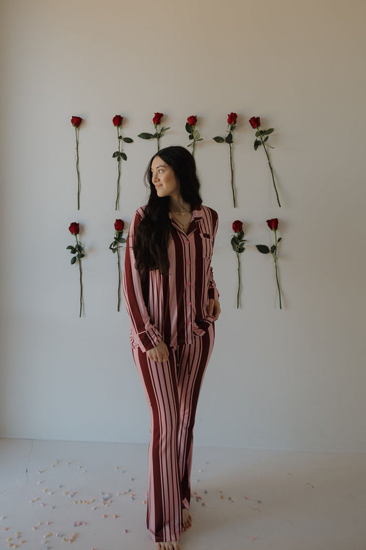 A woman wears the forever french baby Women's Bamboo Button Up Lounge Set in Sweetheart Stripes, standing barefoot by a white wall decorated with red roses and petals scattered on the floor around her.