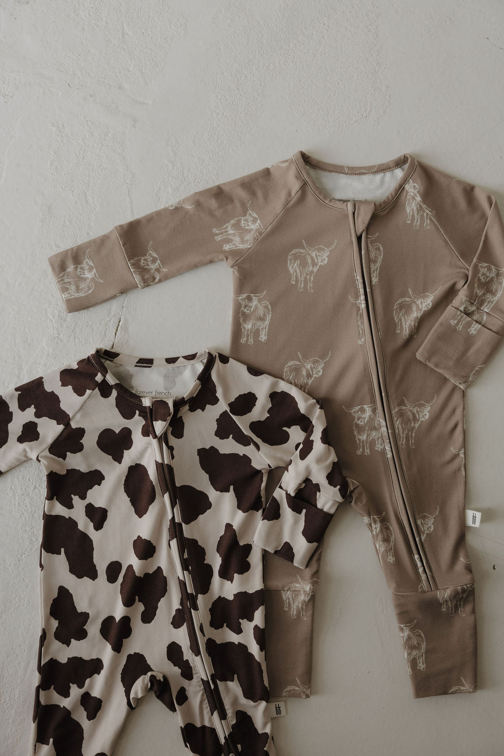 Two Bamboo Zip Pajamas | Moo! by forever french baby rest on a light surface—one with a cow print, the other light brown with white cows. These bamboo long-sleeve onesies have front zippers and are perfect hypoallergenic baby clothing.
