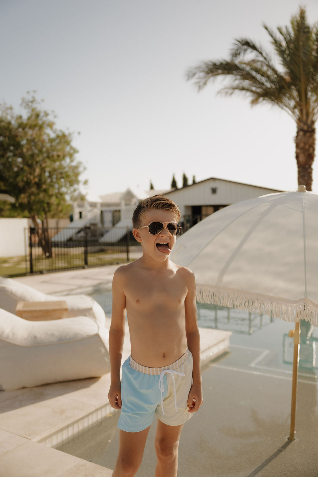 A young boy in lolo webb 2's Child Board Short | Surf & Sand smiles by the pool under bright sunlight, with a white umbrella, outdoor furniture, and palm trees in the background.