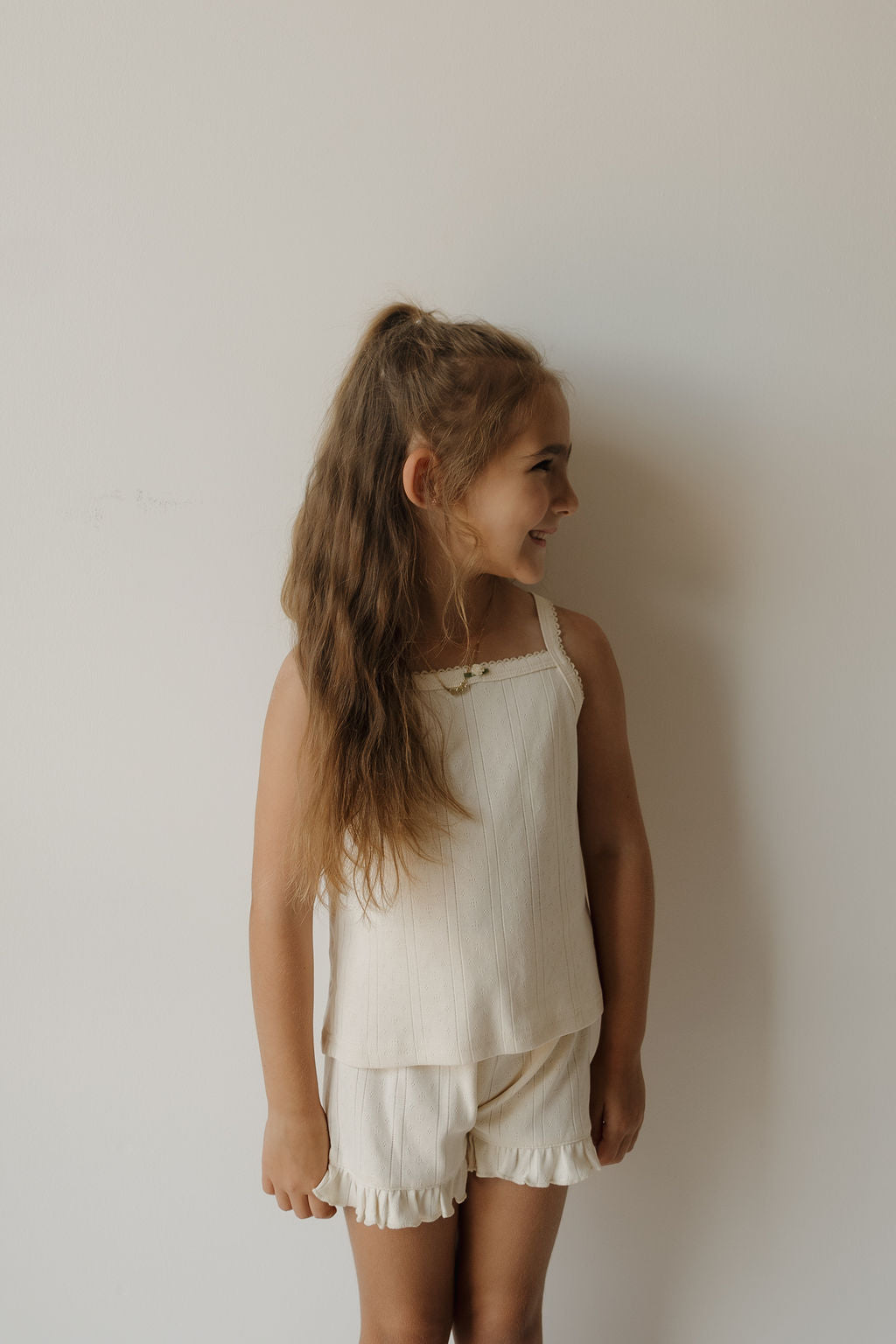 A young girl with long brown hair, dressed in the forever french baby Girl’s Ruffle Short Set | Swan—a cream-colored, sleeveless pointelle top paired with matching ruffle shorts—stands against a plain light-colored wall, smiling and looking to the right.