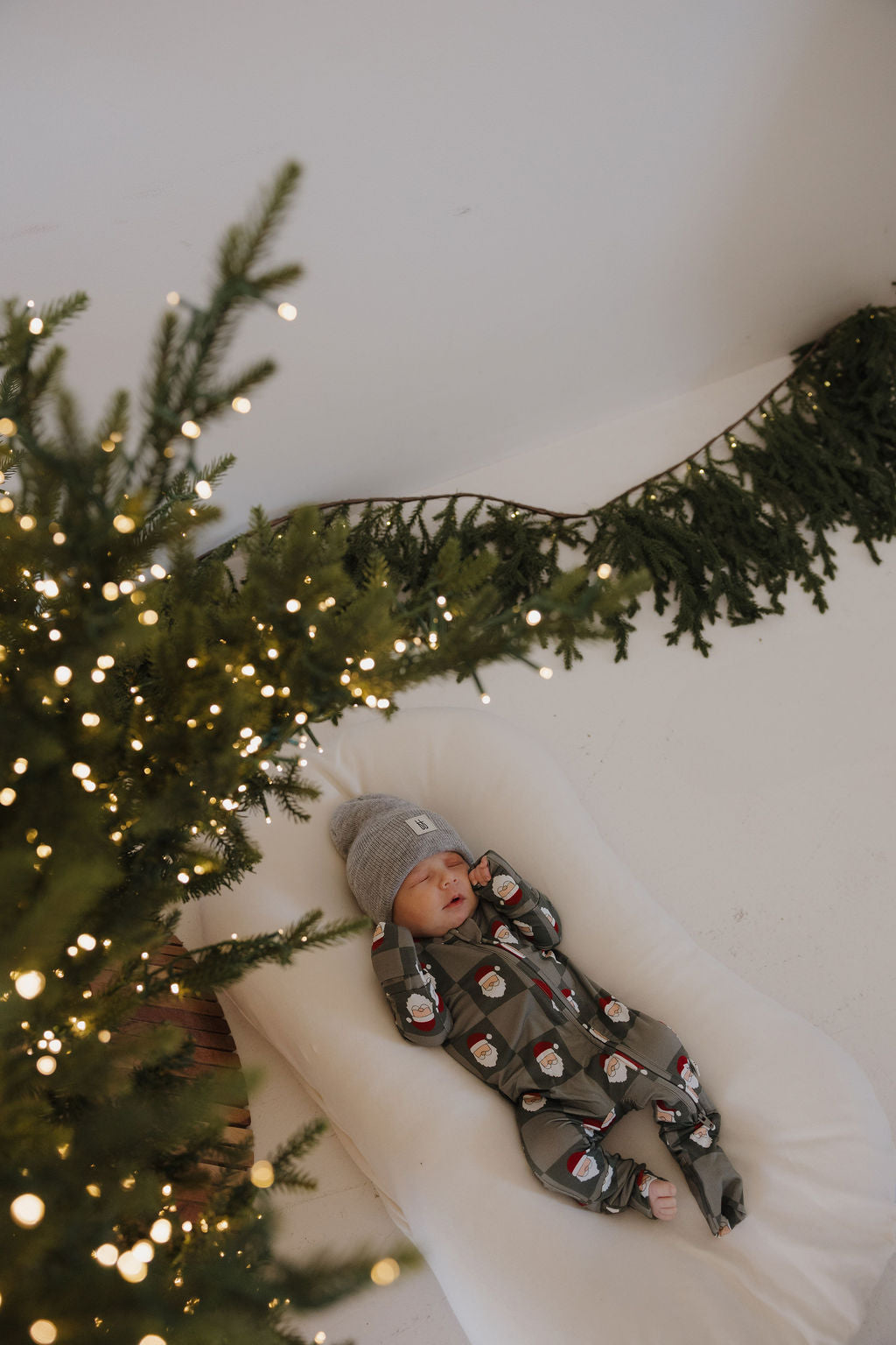 A baby in forever french baby's Bamboo Zip Pajamas | Santa Checkerboard and a gray knit hat lies on a cushioned surface next to a decorated Christmas tree glowing with lights and garland in the background.
