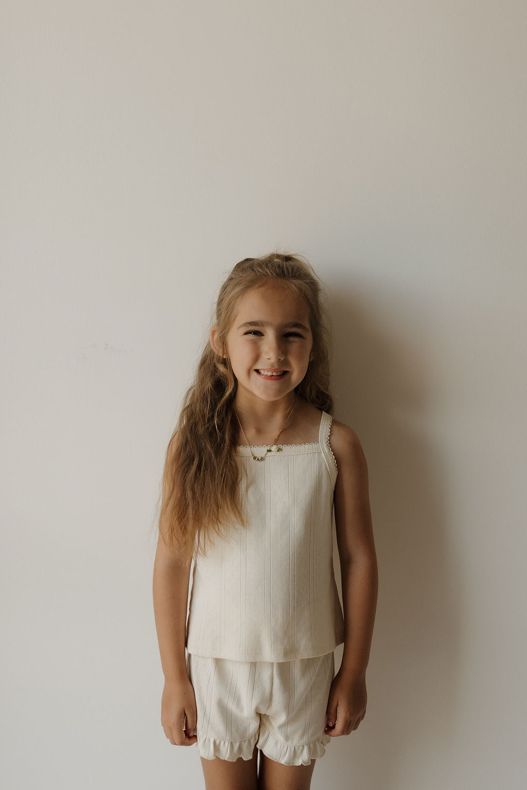 A young girl with long light brown hair stands against a plain white wall, smiling softly as she wears the forever french baby Girl’s Ruffle Short Set | Swan—a sleeveless cream top and matching ruffle shorts made from soft pointelle fabric.