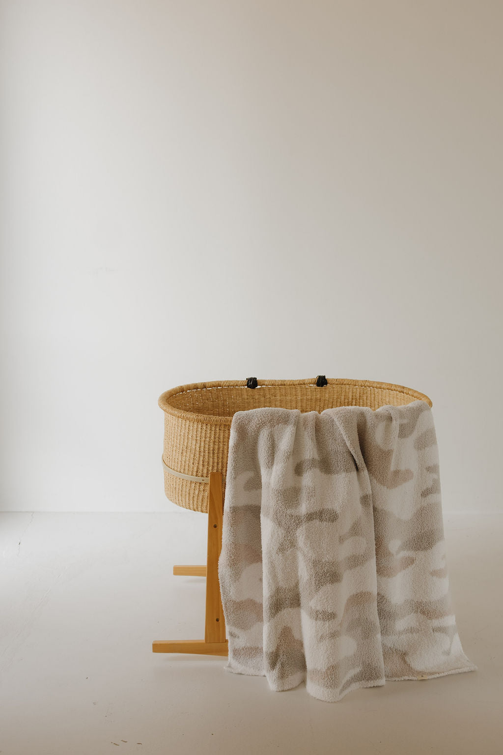 A woven baby bassinet with a wooden stand is in a minimal, light-filled room. A forever french baby Plush Blanket | Camo 2.0, OEKO-TEX certified and soft in white and beige, is draped over the edge of the bassinet.