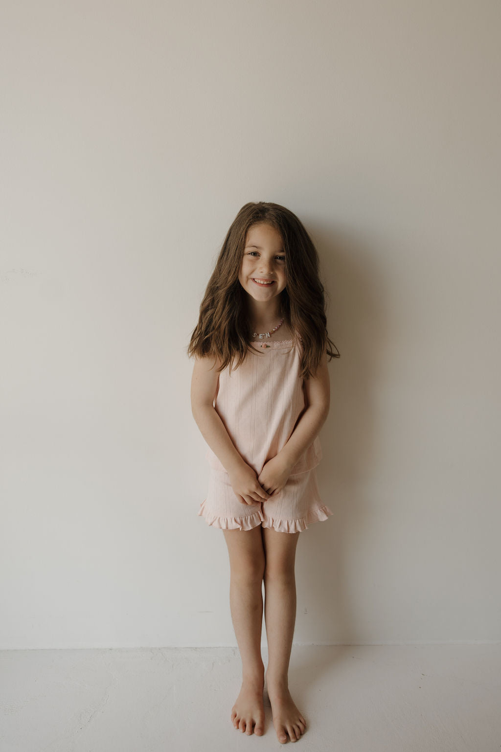 A young girl with long brown hair stands barefoot against a plain white wall, wearing the forever french baby Girl’s Ruffle Short Set in Ballerina—a light pink, sleeveless ensemble crafted from soft 100% cotton. She smiles softly with her hands clasped in front of her, radiating simple charm and comfort.