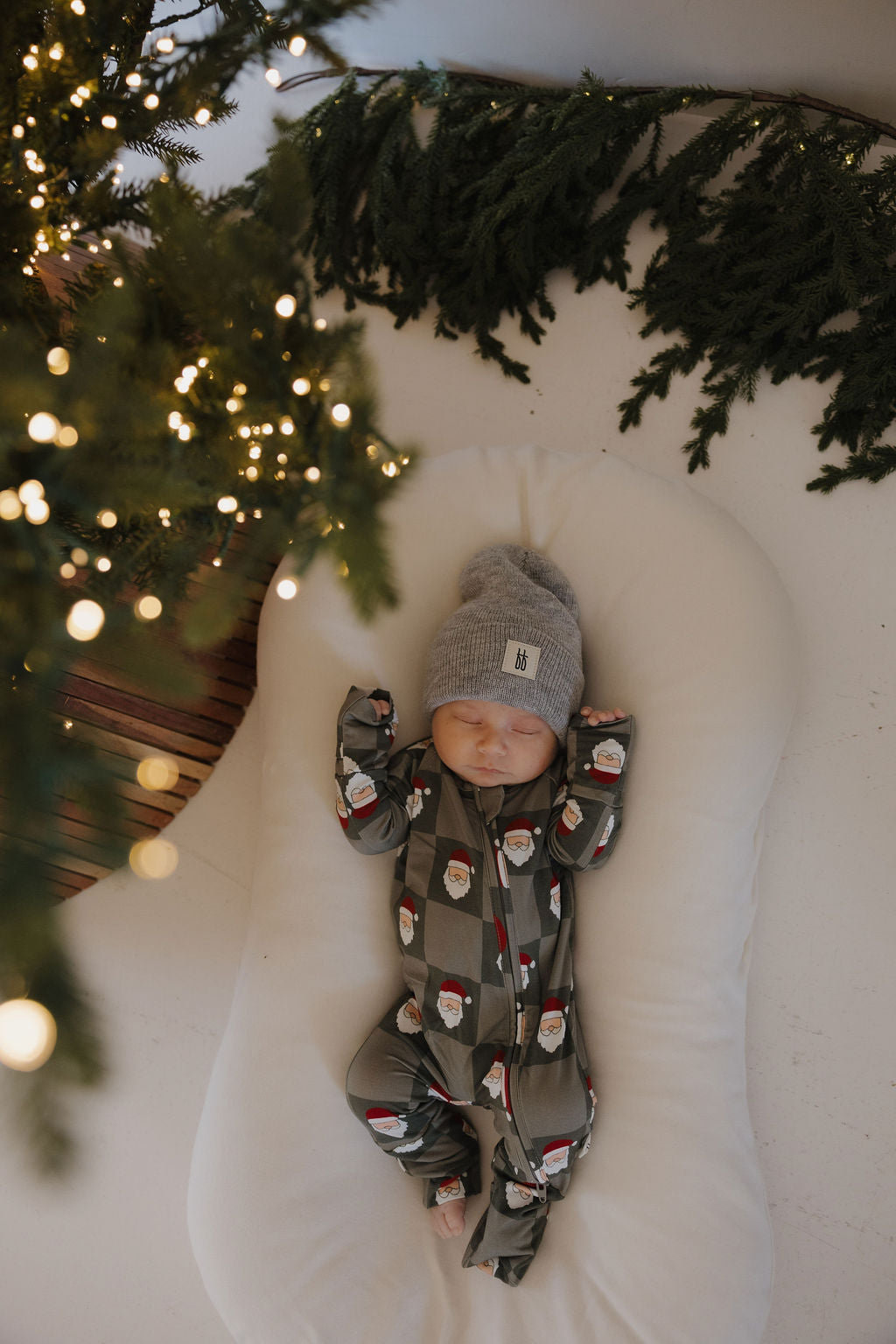 A baby in forever french baby Bamboo Zip Pajamas | Santa Checkerboard and a gray knit hat sleeps peacefully on a white cushion by a Christmas tree, highlighting festive holiday sleepwear.