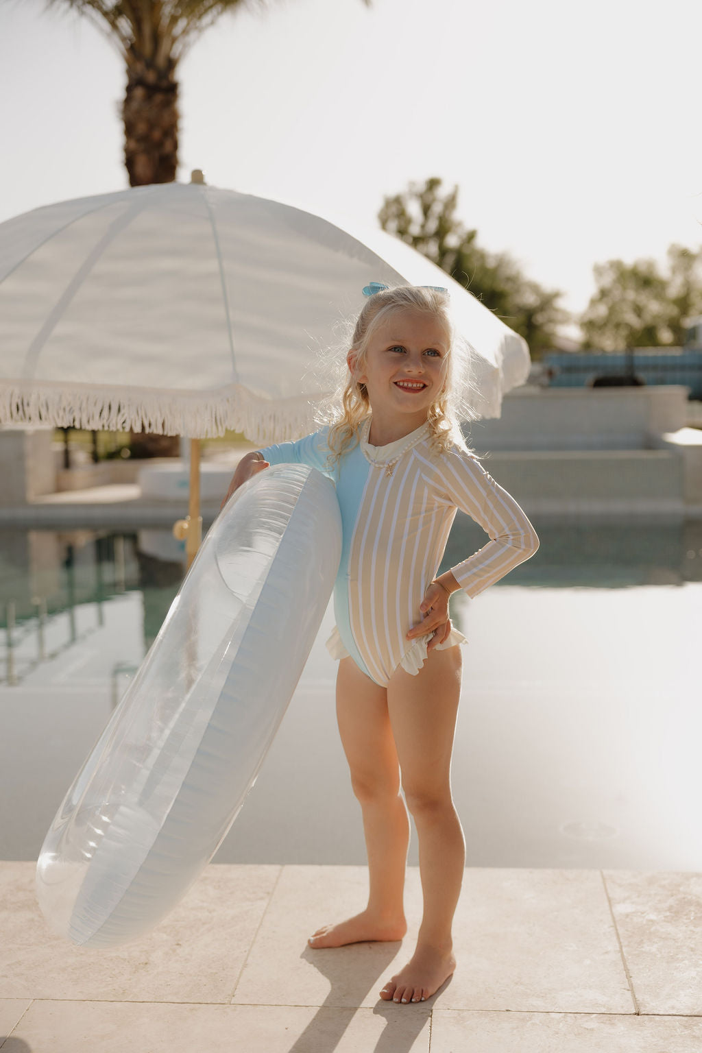 A young girl in the lolo webb 2 Girl Long Sleeve Swimsuit | Surf & Sand smiles by a pool, holding a clear inflatable ring, with a white umbrella and palm tree in the sunny background.