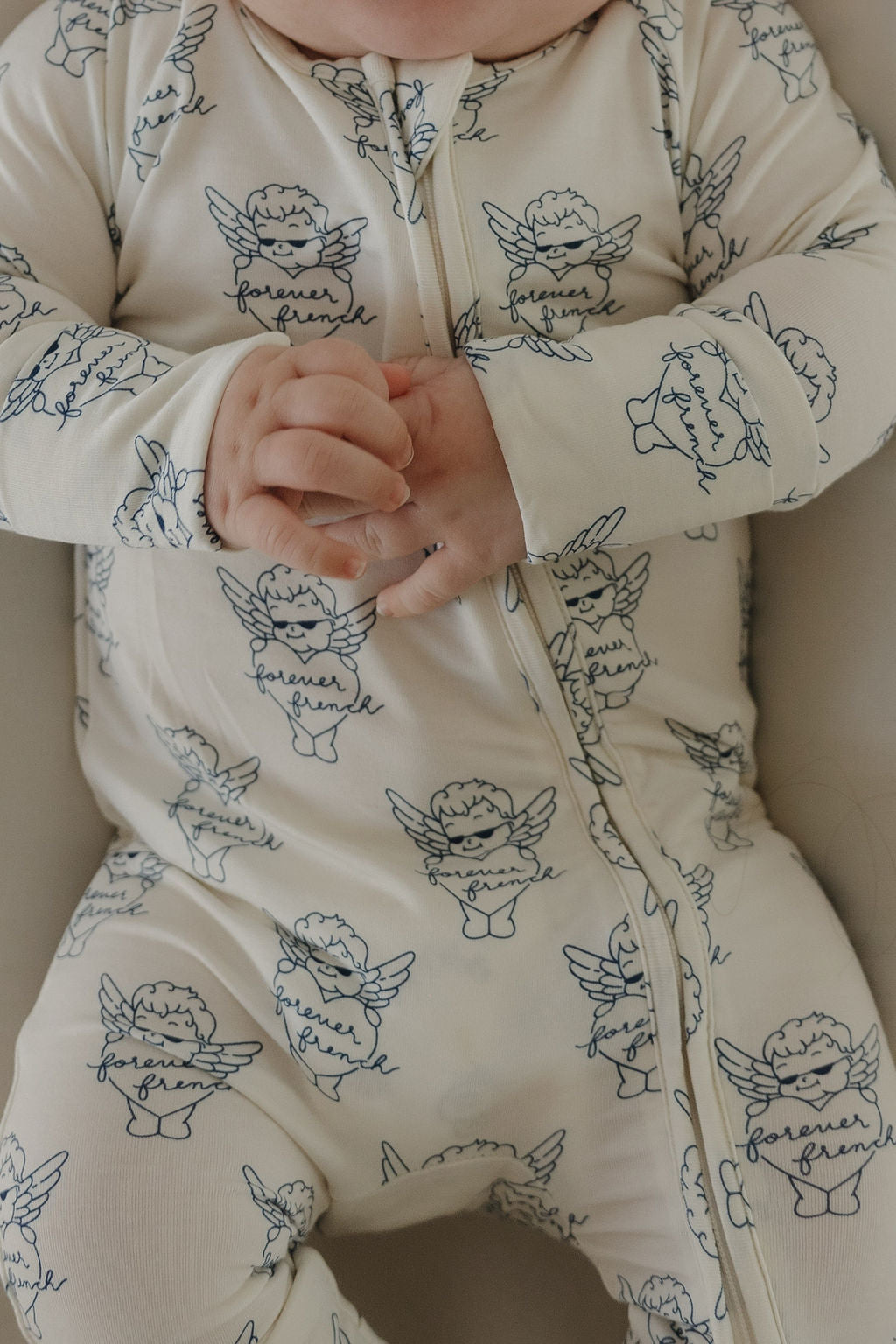 A baby clasps their hands together while wearing forever french baby's cream-colored Bamboo Zip Pajamas from ff Los Angeles, featuring cherubs with "forever friend" banners and hypoallergenic fabric.