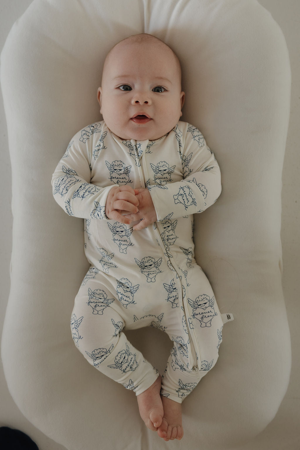 A baby lies on a soft, white cushion wearing forever french baby's Bamboo Zip Pajamas by ff Los Angeles in cream with blue animal print, cozy and calm with hands folded and bare feet peeking out.