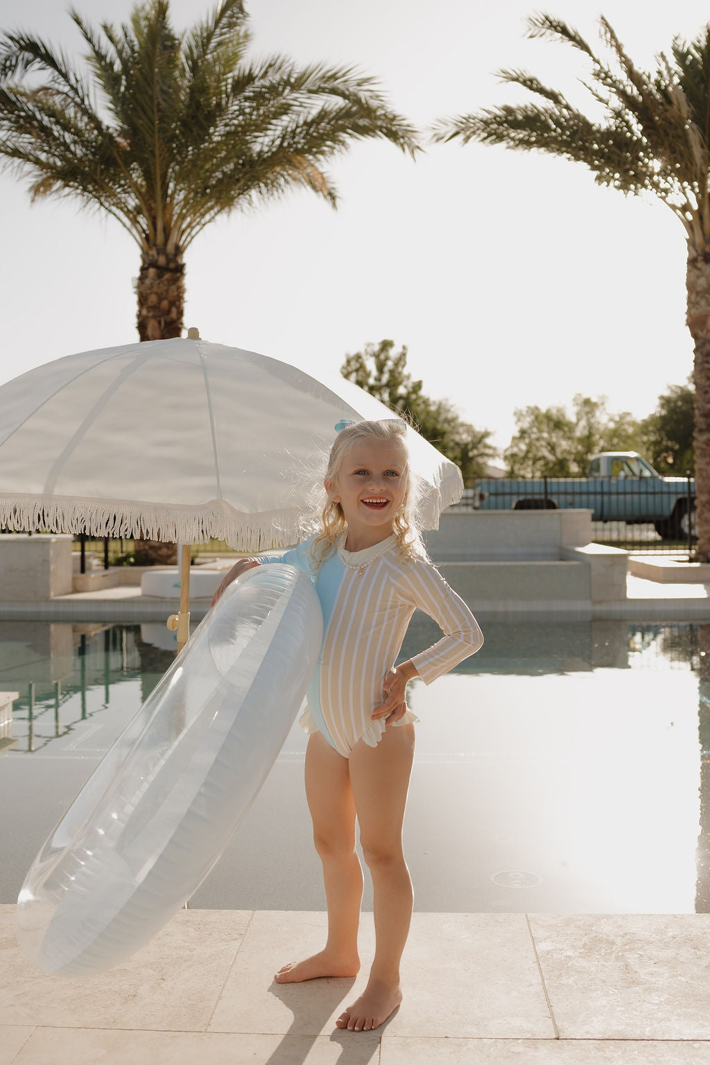 A young girl in the lolo webb 2 Girl Long Sleeve Swimsuit | Surf & Sand stands poolside, smiling with a clear inflatable ring under a white umbrella, palm trees and sunlight in the background.