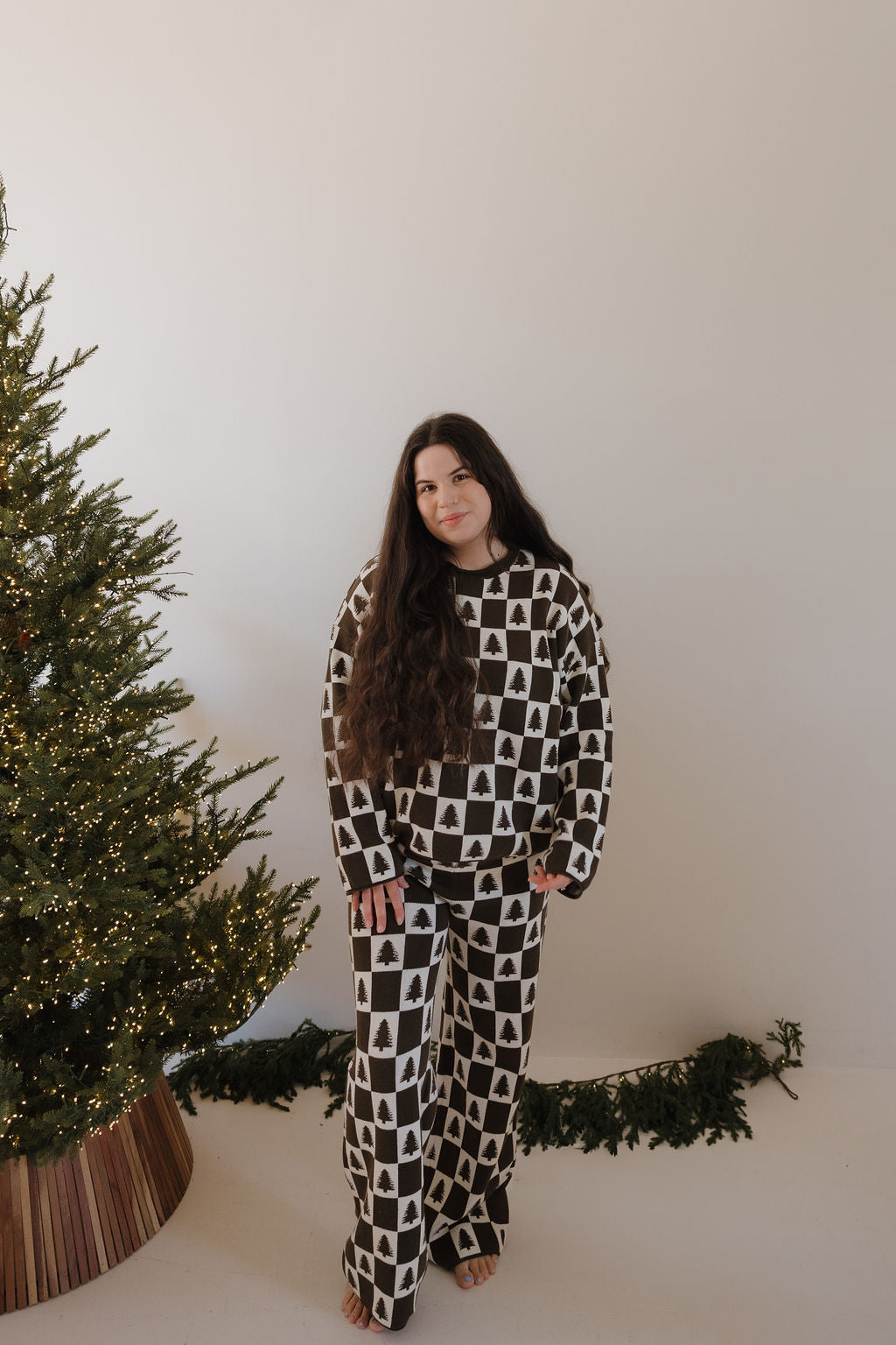 A woman with long brown hair stands barefoot by a decorated Christmas tree, wearing the forever french baby Women's Knit Pant Set in Christmas Tree Checkerboard. The setting is minimal, with some garland on the floor.
