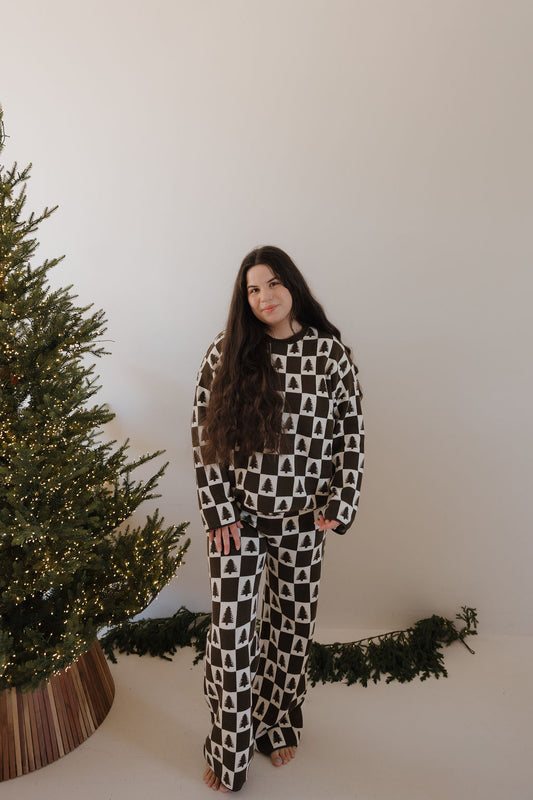 A woman with long brown hair stands barefoot by a decorated Christmas tree, wearing the forever french baby Women's Knit Pant Set in Christmas Tree Checkerboard. The setting is minimal, with some garland on the floor.