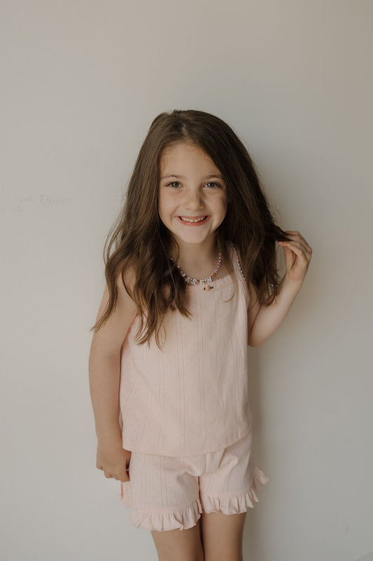 A young girl with long brown hair smiles at the camera as she stands against a plain white wall, wearing the forever french baby Girl’s Ruffle Short Set | Ballerina. This light pink set is crafted from soft 100% cotton pointelle fabric and includes a sleeveless top paired with matching ruffled hem shorts.