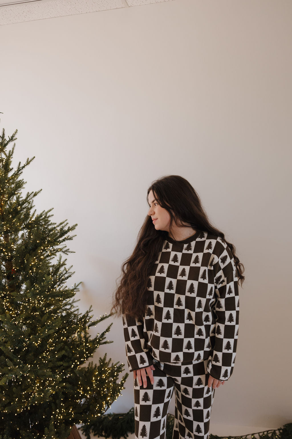 A woman with long brown hair wears the forever french baby Women's Knit Pant Set in the Christmas Tree Checkerboard pattern, standing by a decorated Christmas tree with string lights and looking to her left.