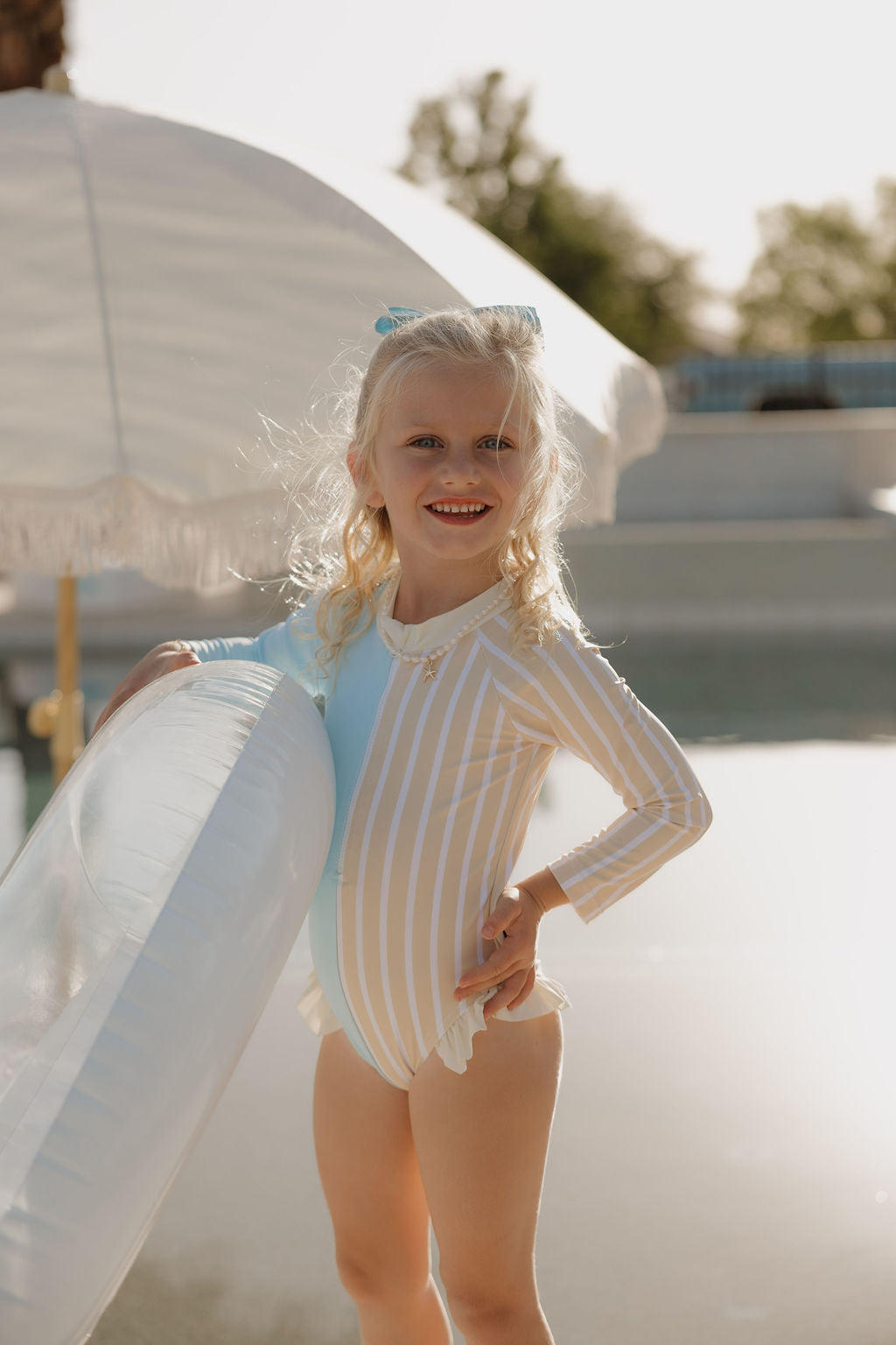 A young girl in the lolo webb 2 Girl Long Sleeve Swimsuit | Surf & Sand smiles by the pool with a white inflatable float, sunlit and playful under a white umbrella, capturing Forever French style.