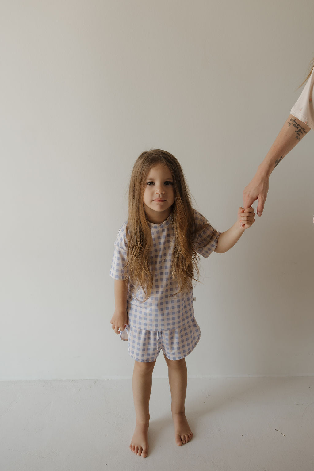 A young child with long hair, barefoot on a white floor and holding an adult’s hand, wears the forever french baby Girl’s Bamboo Short Set | Marilyn against a plain light-colored wall. This hypo-allergenic bamboo short set is gentle on sensitive skin while providing an adorable, stylish look.