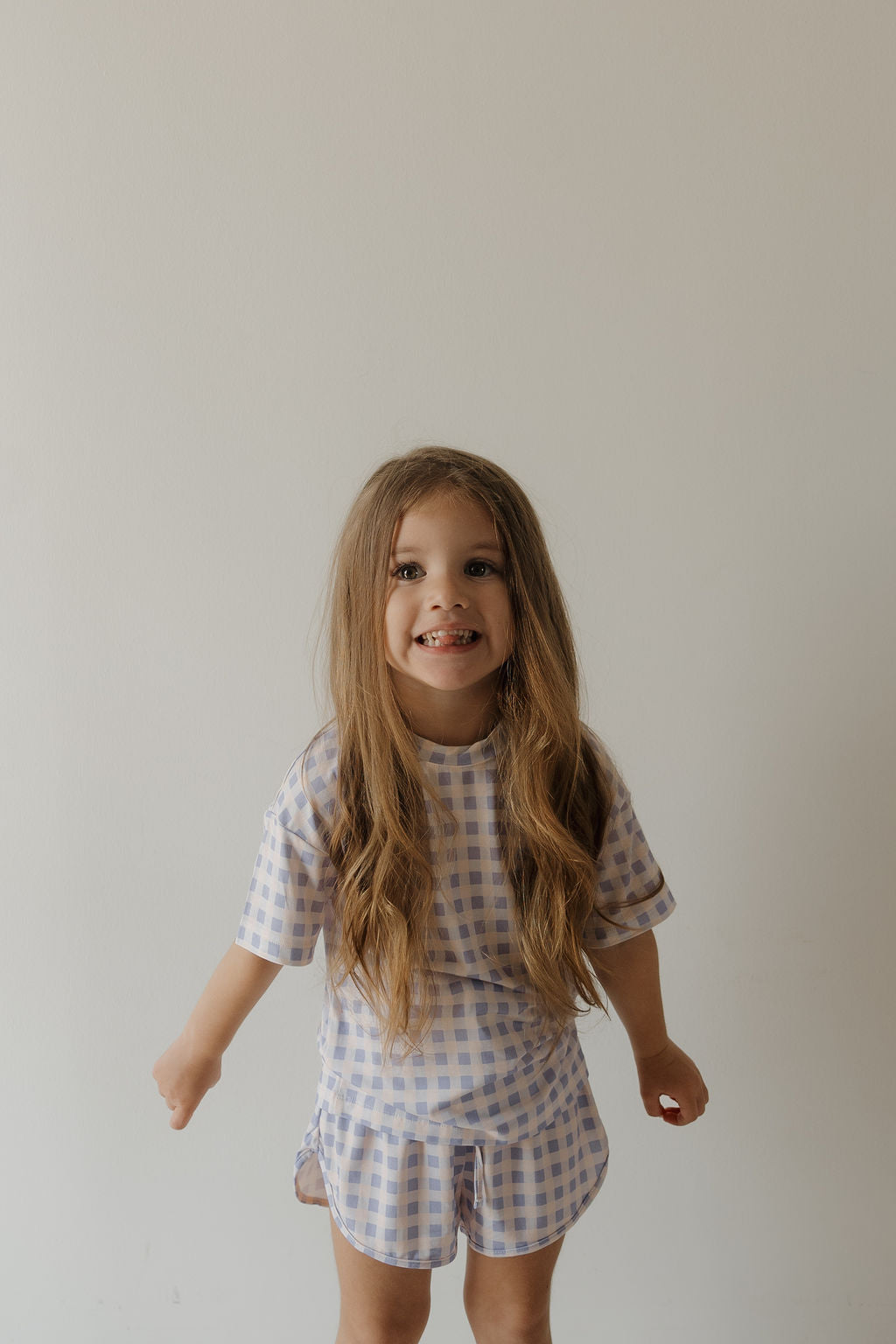 A young girl with long brown hair smiles at the camera, wearing the Girl’s Bamboo Short Set | Marilyn by forever french baby. This light purple and white checkered set, celebrated for its hypo-allergenic properties, stands out against a plain, light-colored wall.