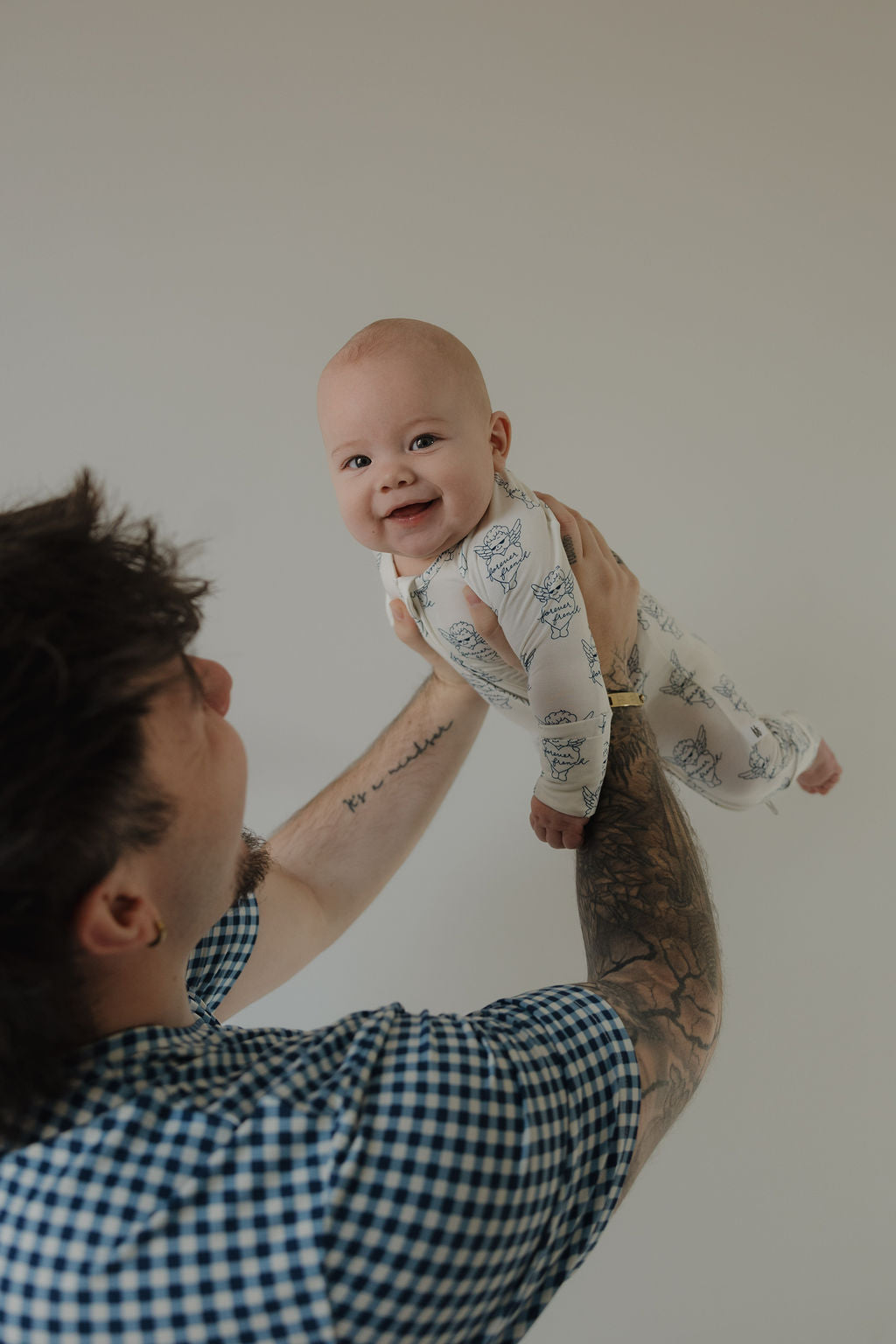 A tattooed person lifts a smiling baby facing the camera, dressed in forever french baby Bamboo Zip Pajamas by ff Los Angeles, against a plain light background.