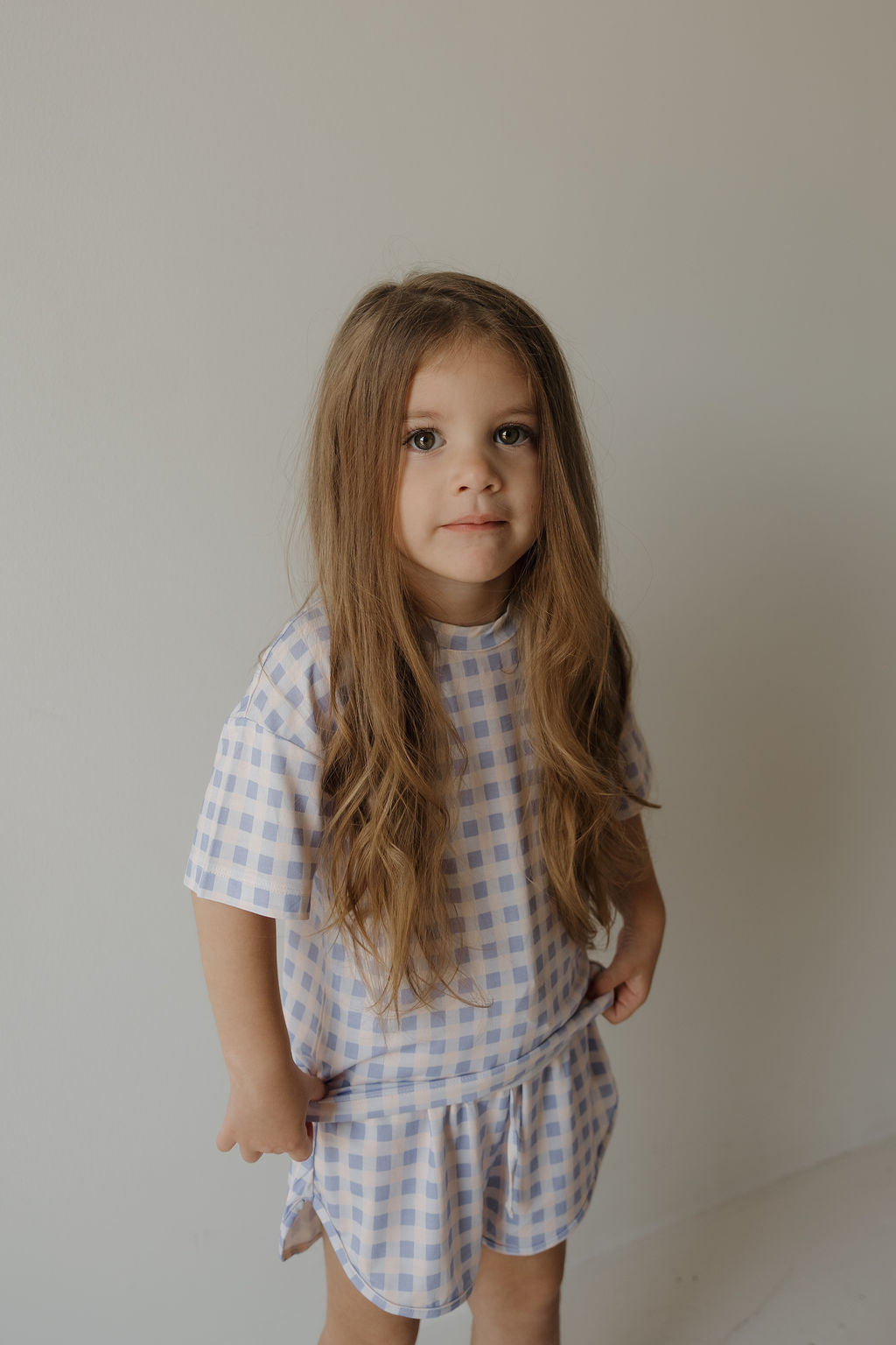 A young child with long brown hair, dressed in the forever french baby Girl’s Bamboo Short Set | Marilyn in blue and white checks, stands against a plain, light-colored wall, gazing slightly past the camera. This hypo-allergenic bamboo set offers exceptional comfort for sensitive skin while delivering a stylish and timeless appearance.