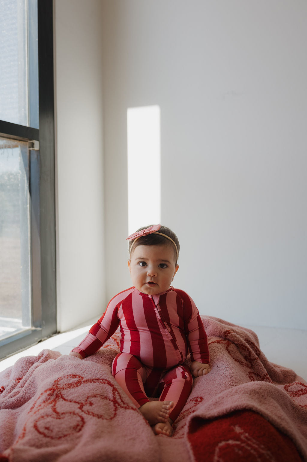 A baby in Minty Cherries Bamboo Zip Pajamas | Iconic Stripe—a red and pink striped outfit with a matching headband—sits on a pink blanket by a sunlit window, looking toward the camera.