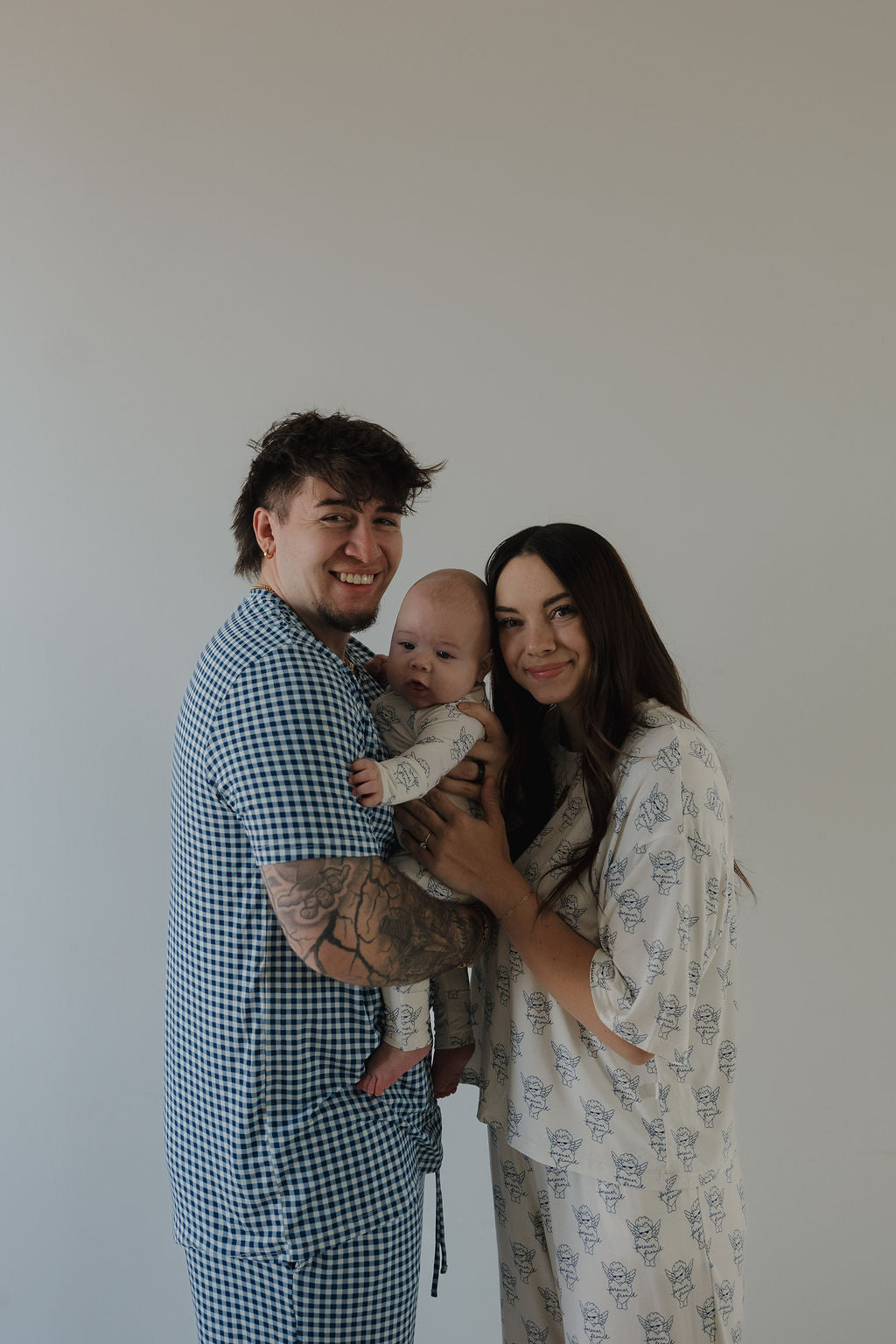 A smiling couple stands together holding a baby in hypoallergenic clothing. The man wears blue checkered pajamas with arm tattoos, while the woman wears Bamboo Zip Pajamas by forever french baby. The background is plain and light-colored.