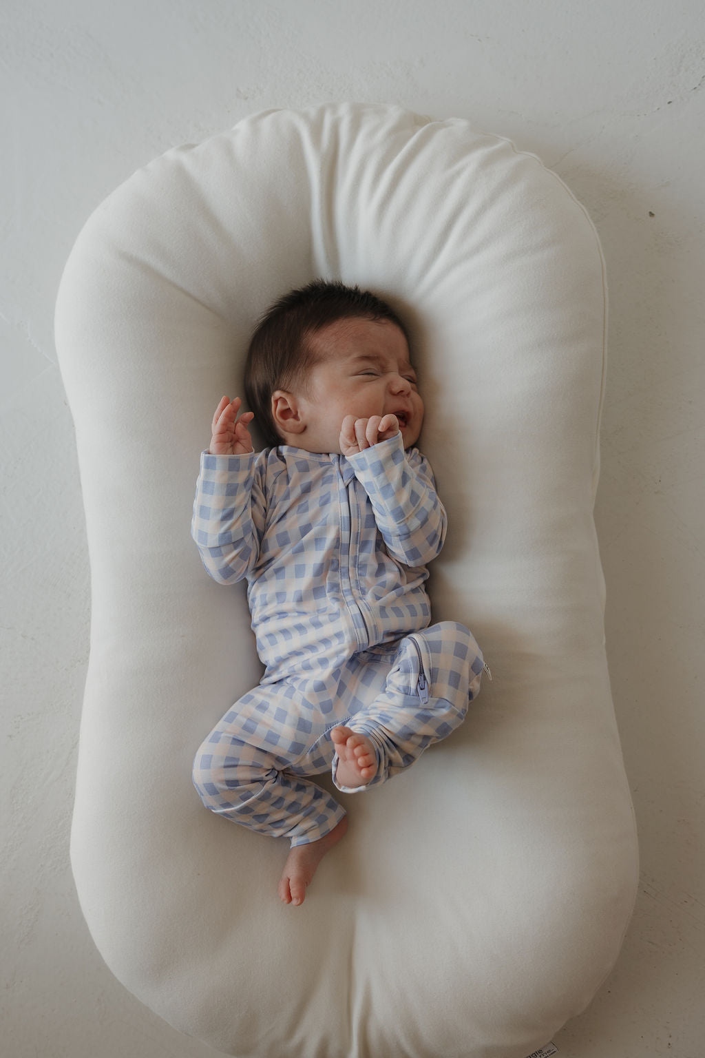 A baby dressed in the Marilyn Bamboo Zip Pajamas from forever french baby, featuring a blue and white checkered pattern, lies on a white cushioned pad with eyes closed and one hand near their face, appearing to cry or fuss. The hypoallergenic, breathable design ensures comfort for sensitive skin.