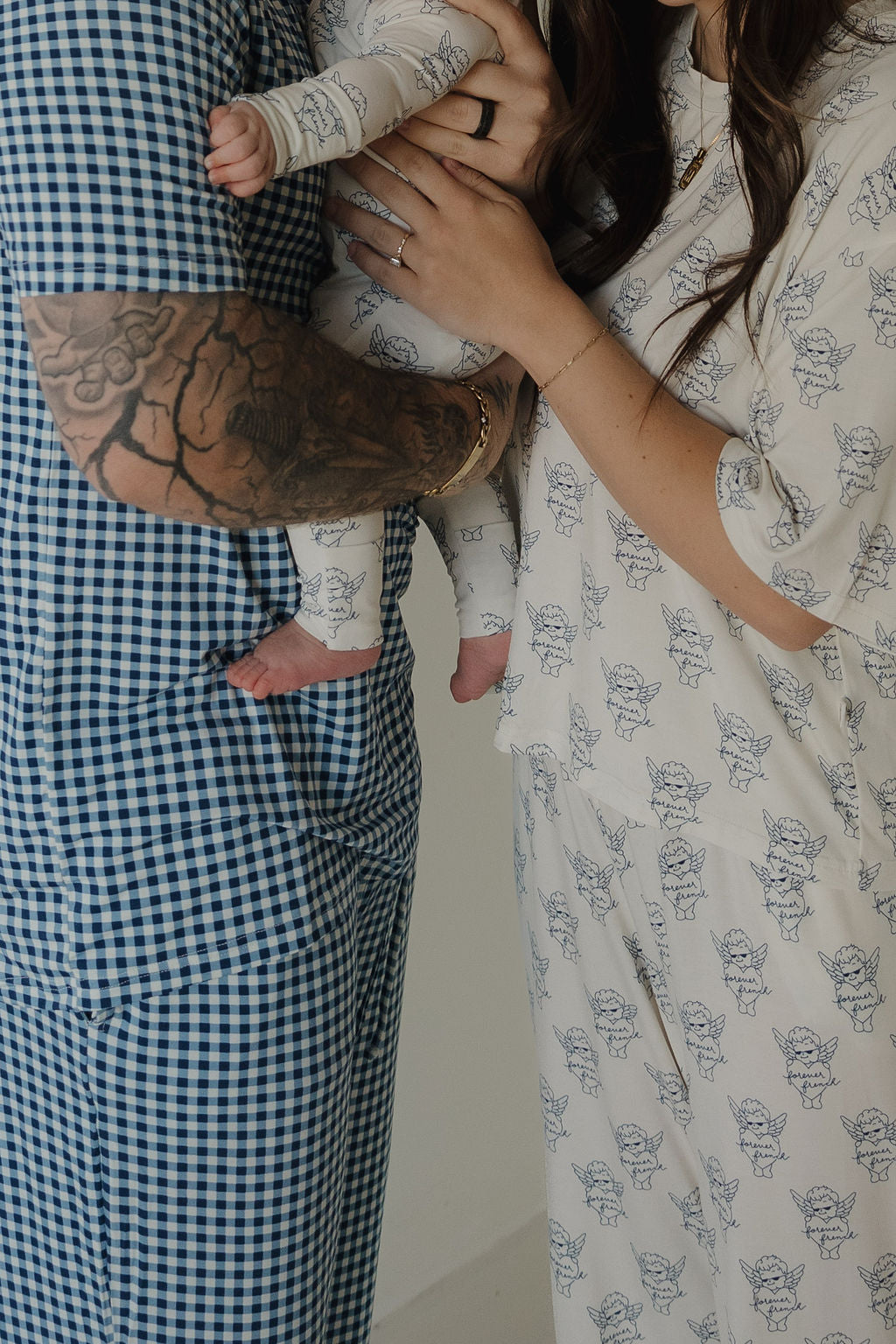 A tattooed man in a blue checkered outfit holds a baby wearing forever french baby Bamboo Zip Pajamas by ff Los Angeles, standing beside a woman in a matching printed dress, all facing each other with arms gently wrapped around the baby.