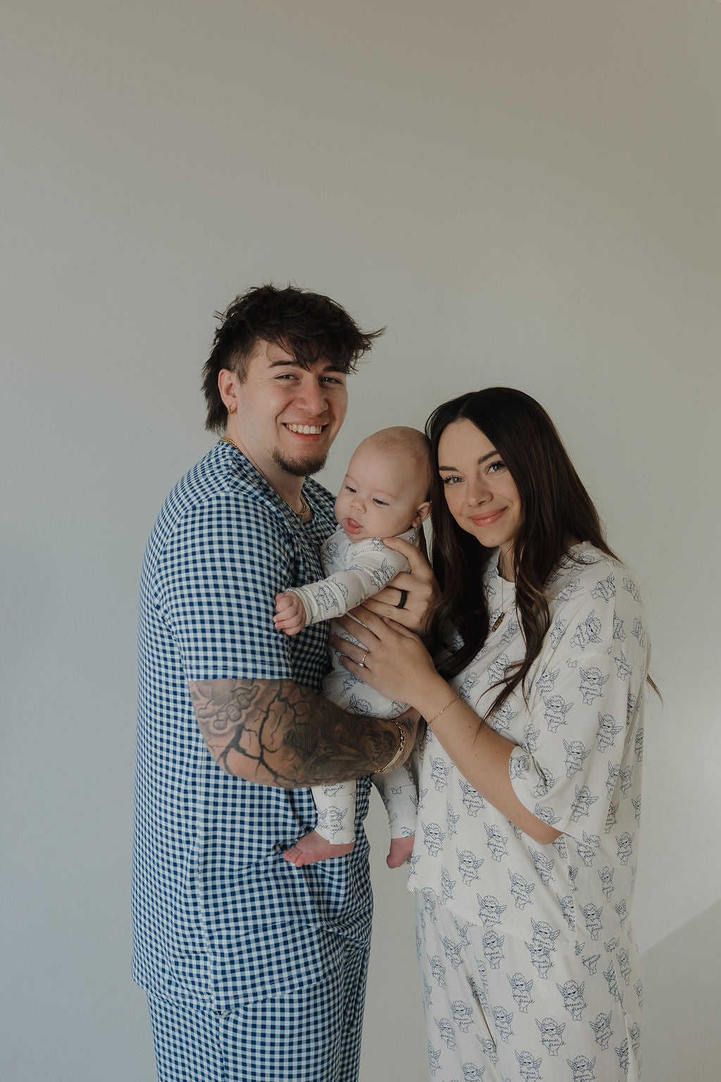 A smiling couple stands together holding a baby dressed in forever french baby’s Bamboo Zip Pajamas by ff Los Angeles. The man wears blue checkered pajamas and has tattooed arms; the woman wears white pajamas with a blue pattern.