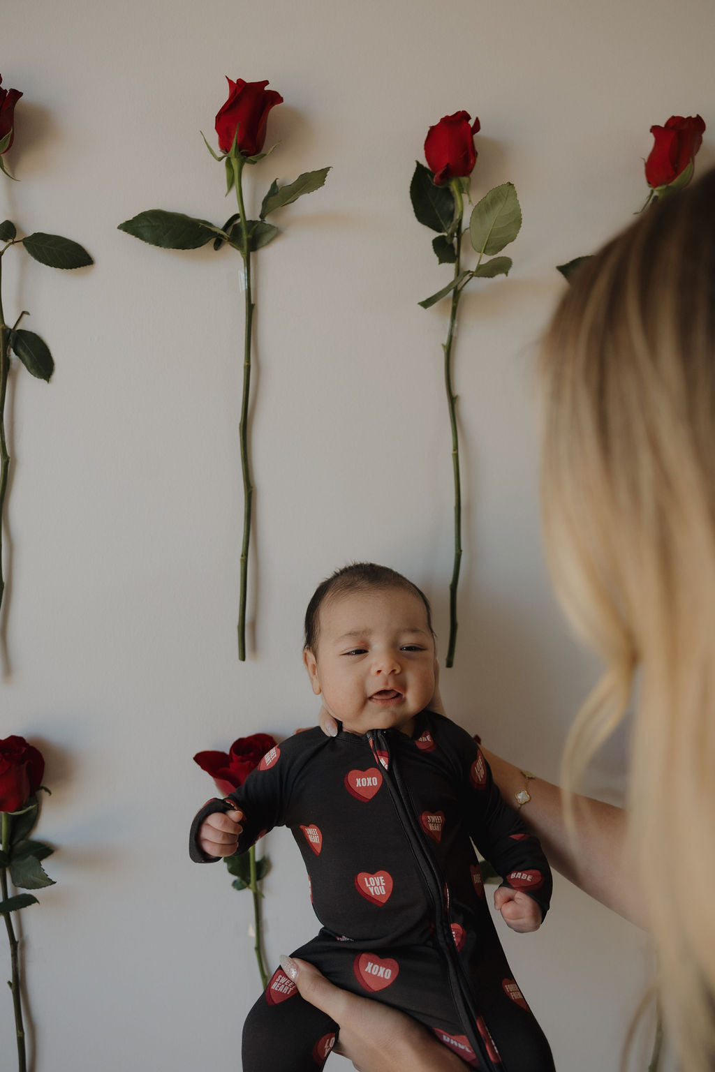 A baby in forever french baby's Bamboo Zip Pajamas | Love Notes – Charcoal, featuring red hearts on black, is held by a blonde adult. Red roses are attached vertically to the light-colored wall behind them.