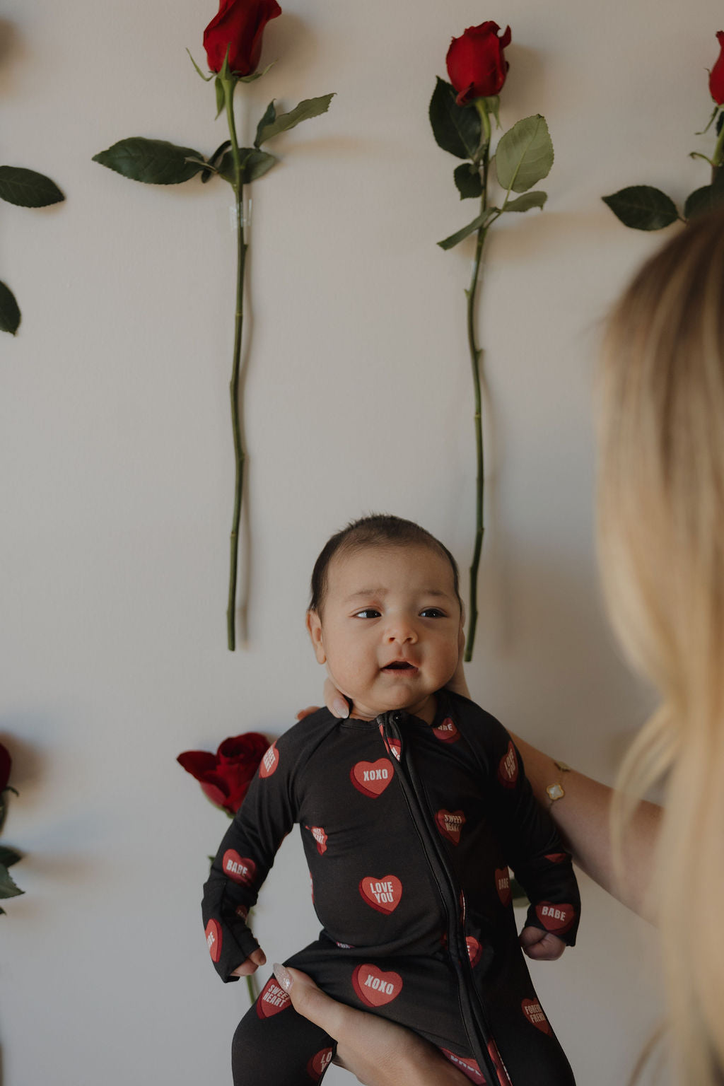 A baby in forever french baby's Bamboo Zip Pajamas | Love Notes – Charcoal sits upright, supported by an adult. Red roses are displayed vertically on a light-colored wall in the background.