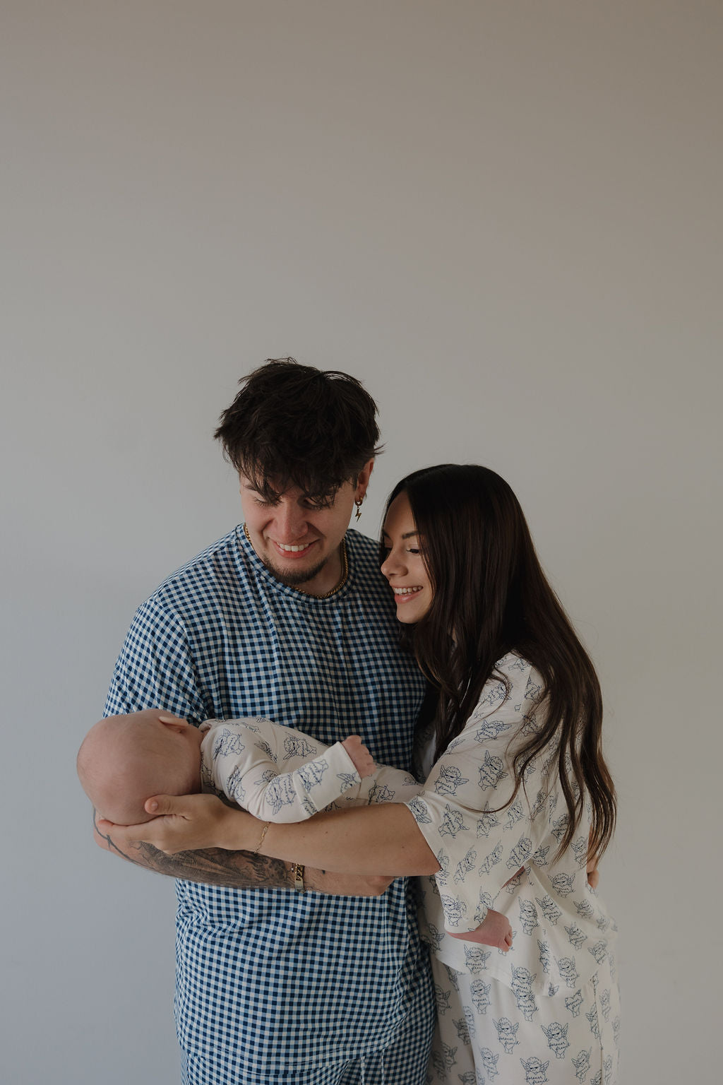 A man and woman smile at each other while holding a baby in Bamboo Zip Pajamas from forever french baby. A plain, light background highlights the happy family enjoying hypoallergenic sleepwear by ff Los Angeles.