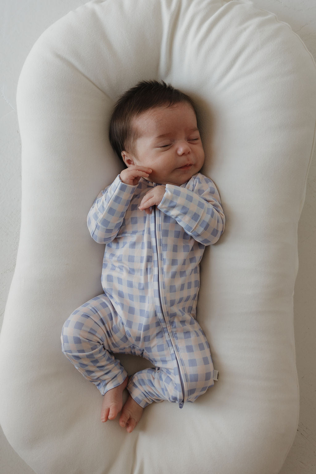 A baby with dark hair sleeps peacefully on a white cushioned lounger, wearing forever french baby’s Bamboo Zip Pajamas in the Marilyn style—a blue and white checkered zip-up onesie made from breathable bamboo fabric to ensure comfort all night long.