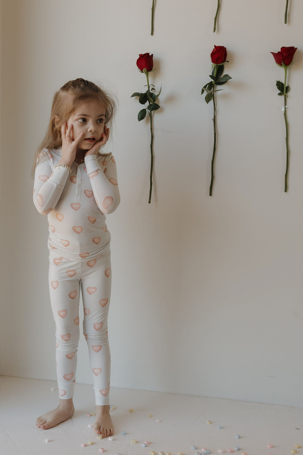 A young girl in forever french baby Bamboo Two Piece Pajamas | Love Notes - Grey stands barefoot among flower petals, touching her face and looking aside, with three red roses attached vertically to the wall behind her.