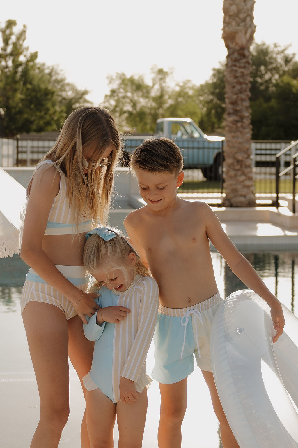 Three kids in swimsuits stand by a pool. The younger girl wears a lolo webb 2 Girl Long Sleeve Swimsuit | Surf & Sand in Sun Rays print. A white pool float, trees, and a parked truck are visible in the background.