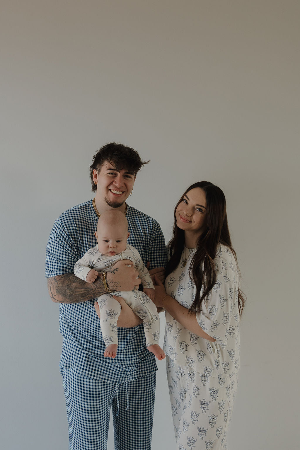 A smiling man in a blue check lounge set holds a baby in printed pajamas, standing next to a woman wearing the Women's Boxy Tee Lounge Set by forever french baby. They pose together before a plain, light-colored background.
