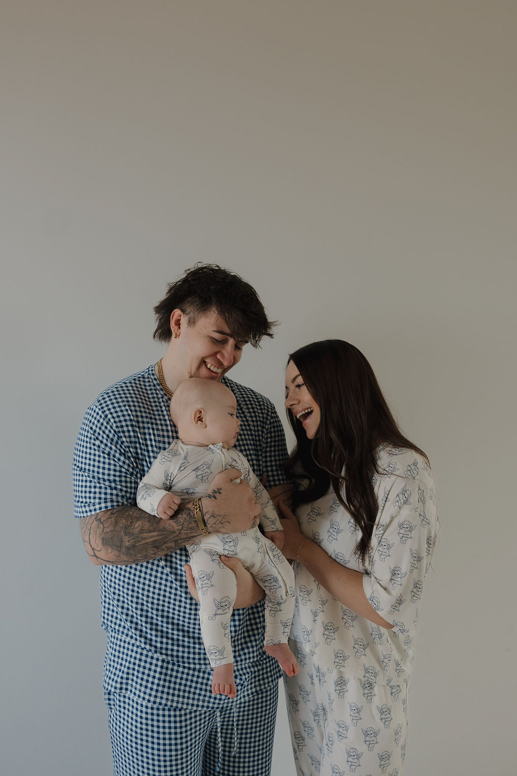A smiling man holds a baby beside a woman; all three happily look at each other while wearing matching forever french baby Bamboo Zip Pajamas by ff Los Angeles, standing against a plain background.