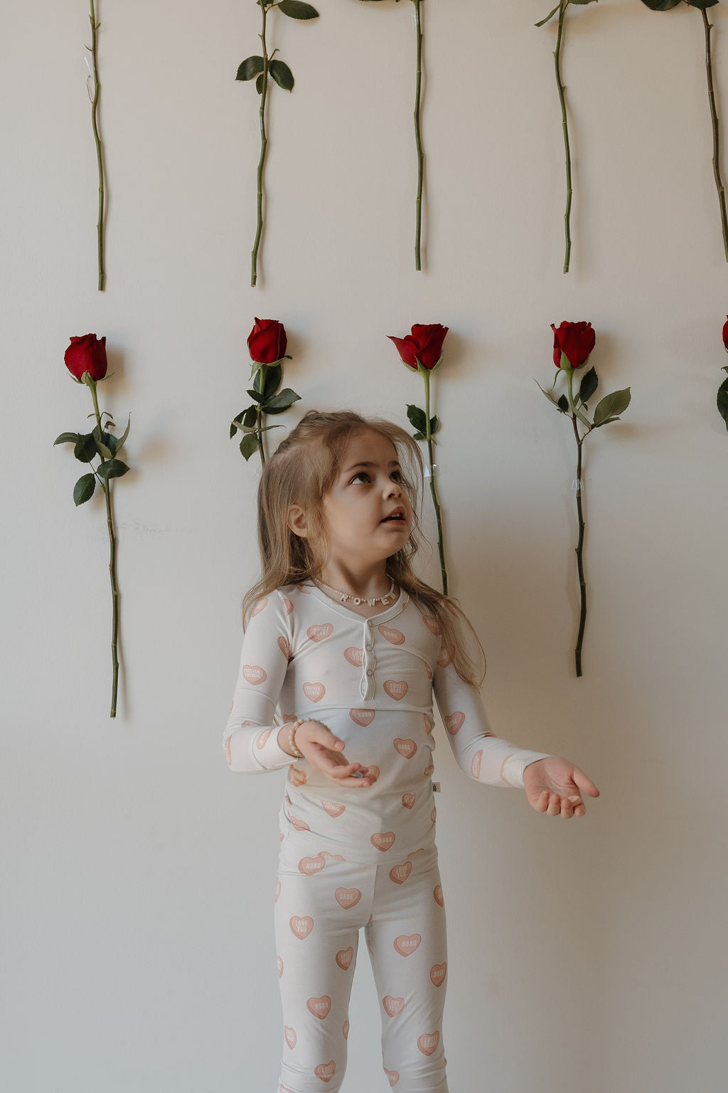 A young girl in forever french baby’s Bamboo Two Piece Pajamas | Love Notes - Grey stands before a white wall with red roses, gazing upward—showcasing breathable kids’ sleepwear ideal for sweet dreams.