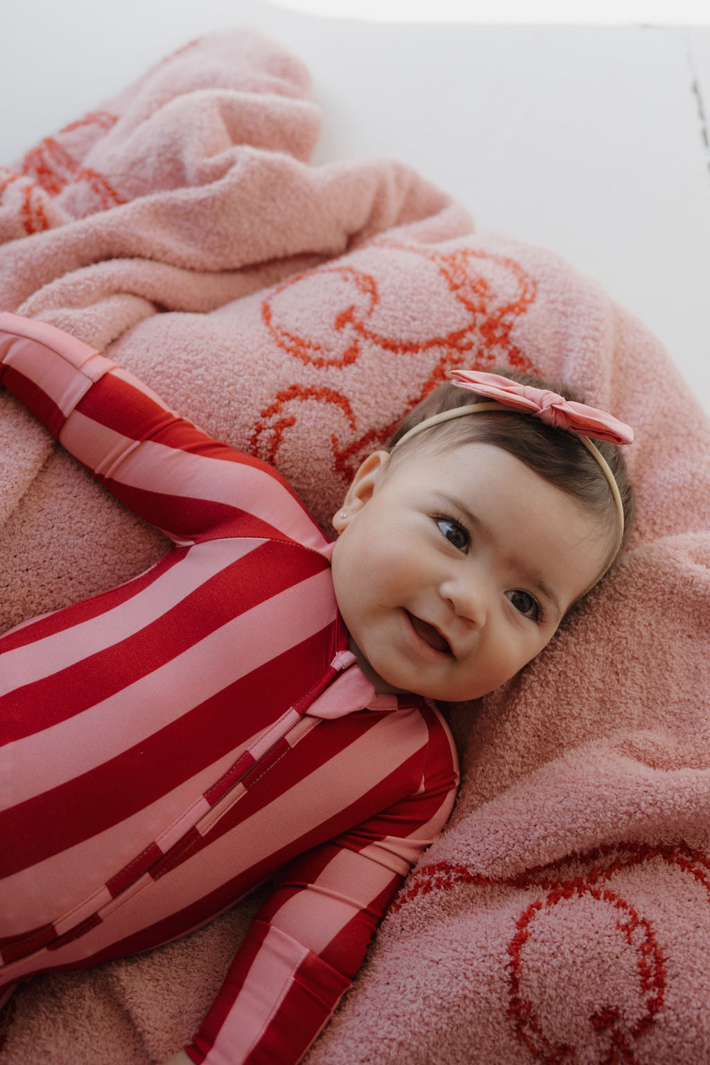 A baby in Minty Cherries Bamboo Zip Pajamas | Iconic Stripe, featuring pink and red stripes, lies on a soft pink blanket with a matching headband, smiling gently.
