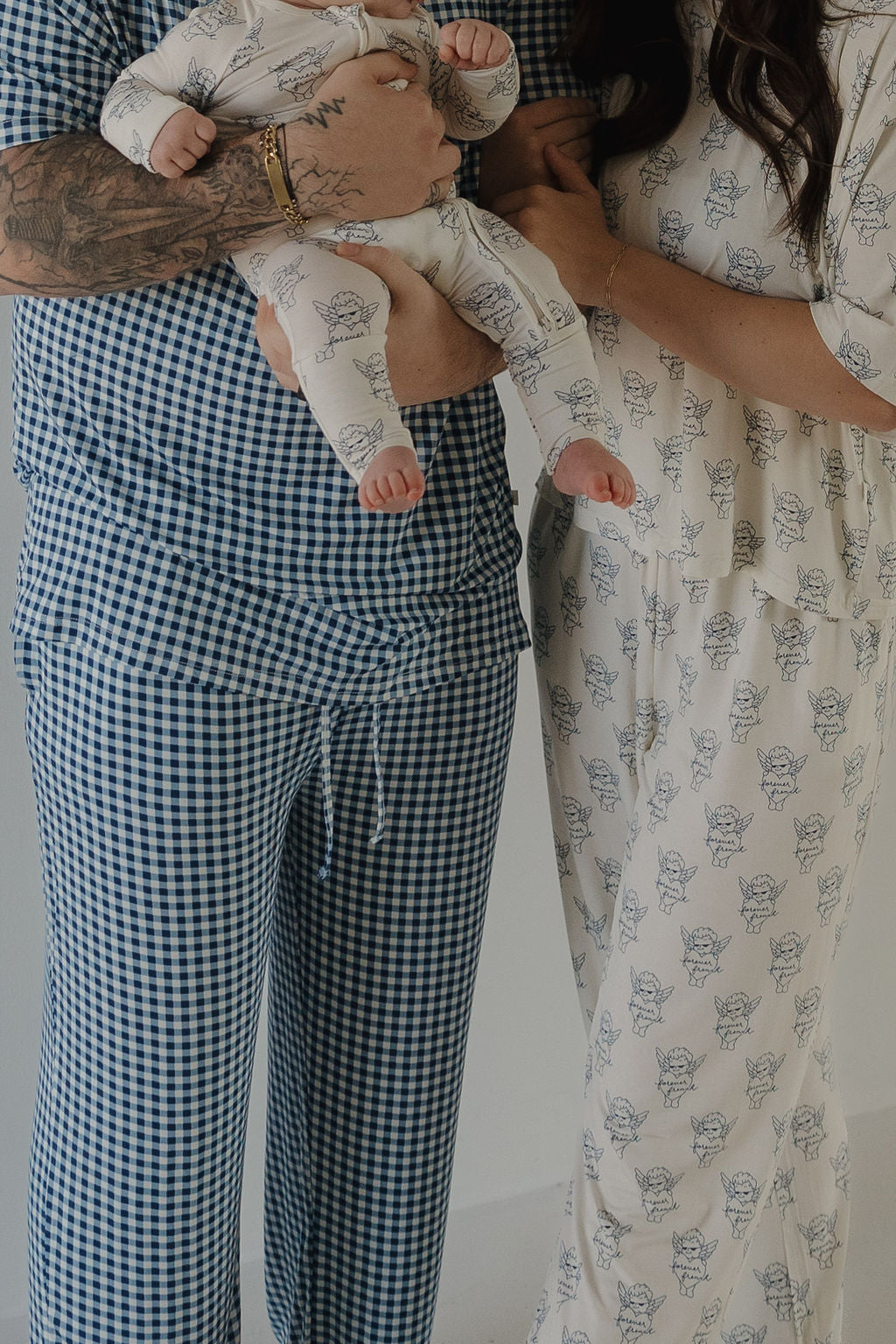 A man in forever french baby Men's Short Sleeve Pajamas | Blue Gingham holds a baby in hypoallergenic animal print pajamas, standing next to a woman in matching bamboo sleepwear. Only their bodies and part of their faces are visible.