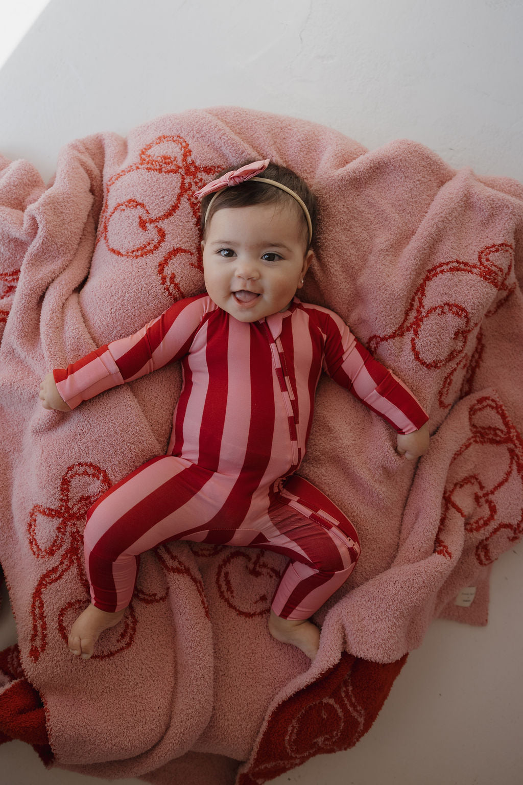 A baby in Minty Cherries Bamboo Zip Pajamas, Iconic Stripe design, and matching headband smiles while lying on a pink blanket with red patterns.