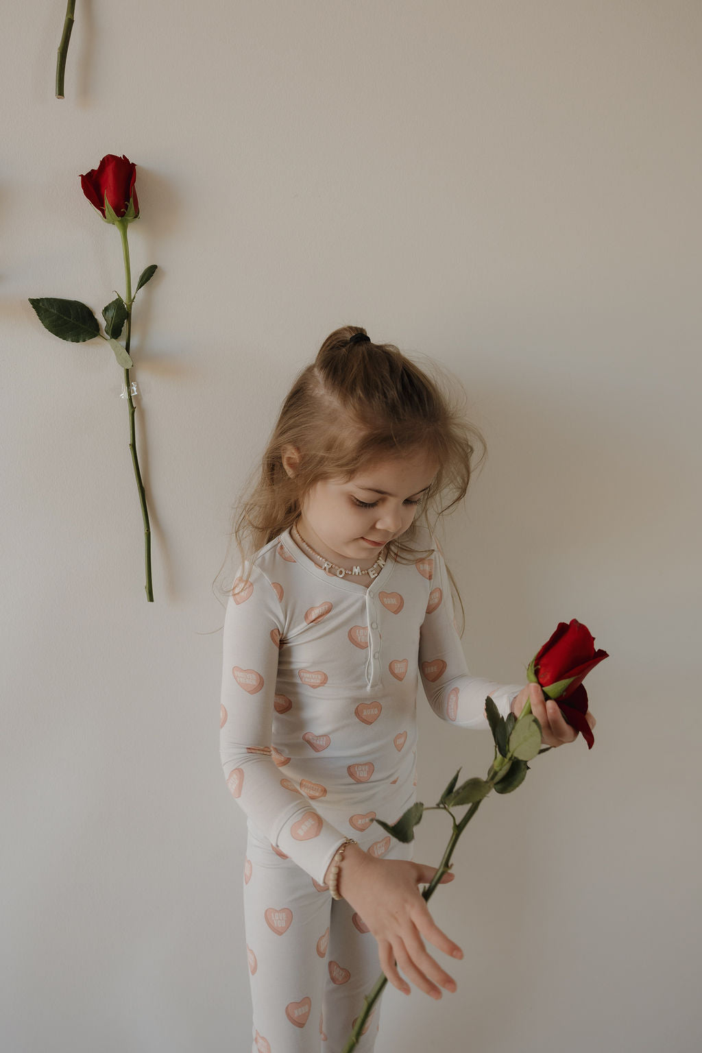 A young girl in forever french baby’s Bamboo Two Piece Pajamas | Love Notes - Grey holds a red rose, standing by a plain wall decorated with two more roses, as she gazes thoughtfully at the flower in her hands.