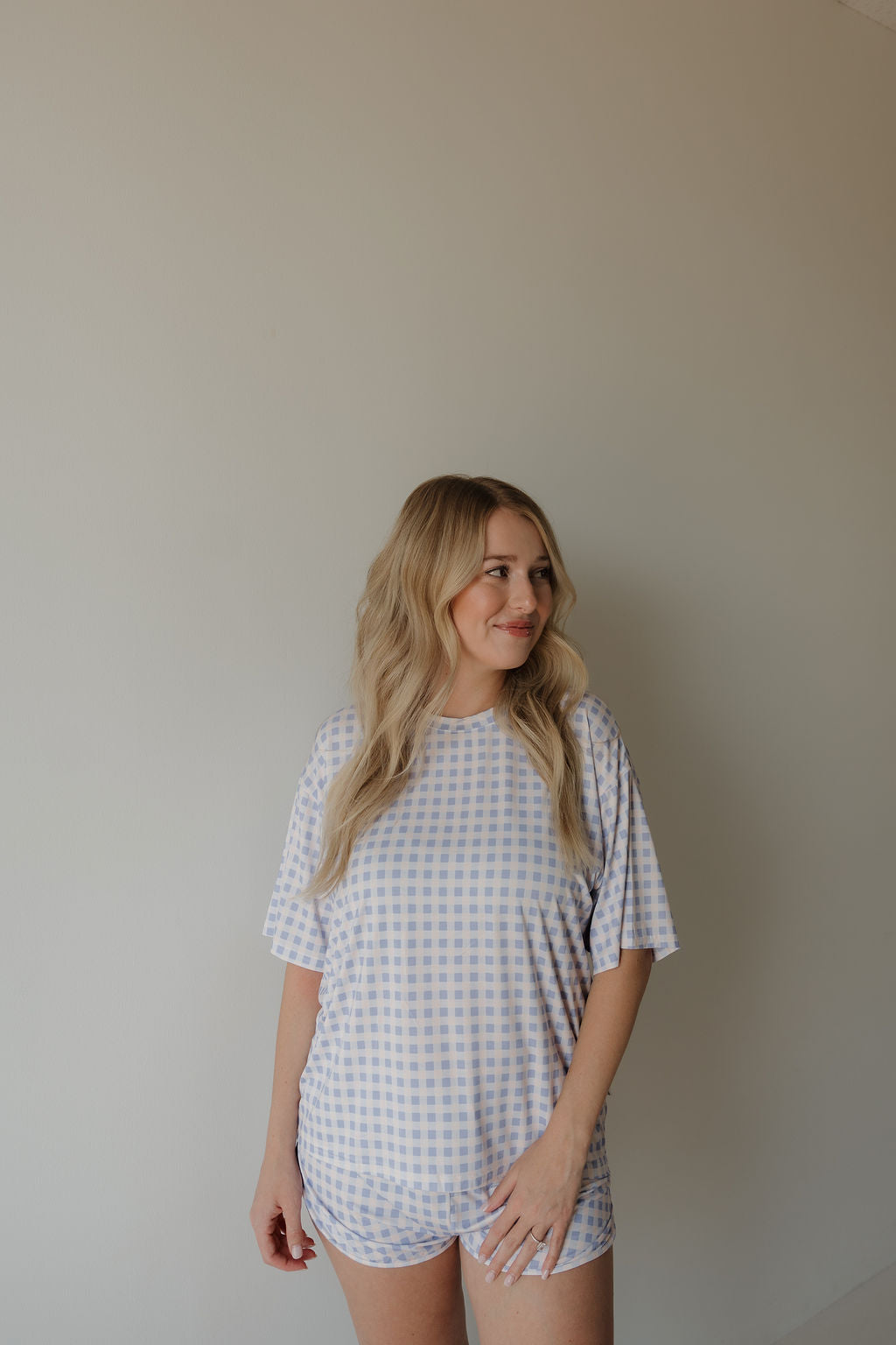 A woman with long, wavy blonde hair wears the forever french baby Women’s Bamboo Short Set | Marilyn—a light blue and white checkered, hypo-allergenic loungewear pajama set—while standing against a plain, light-colored wall and smiling slightly as she looks off to the side.