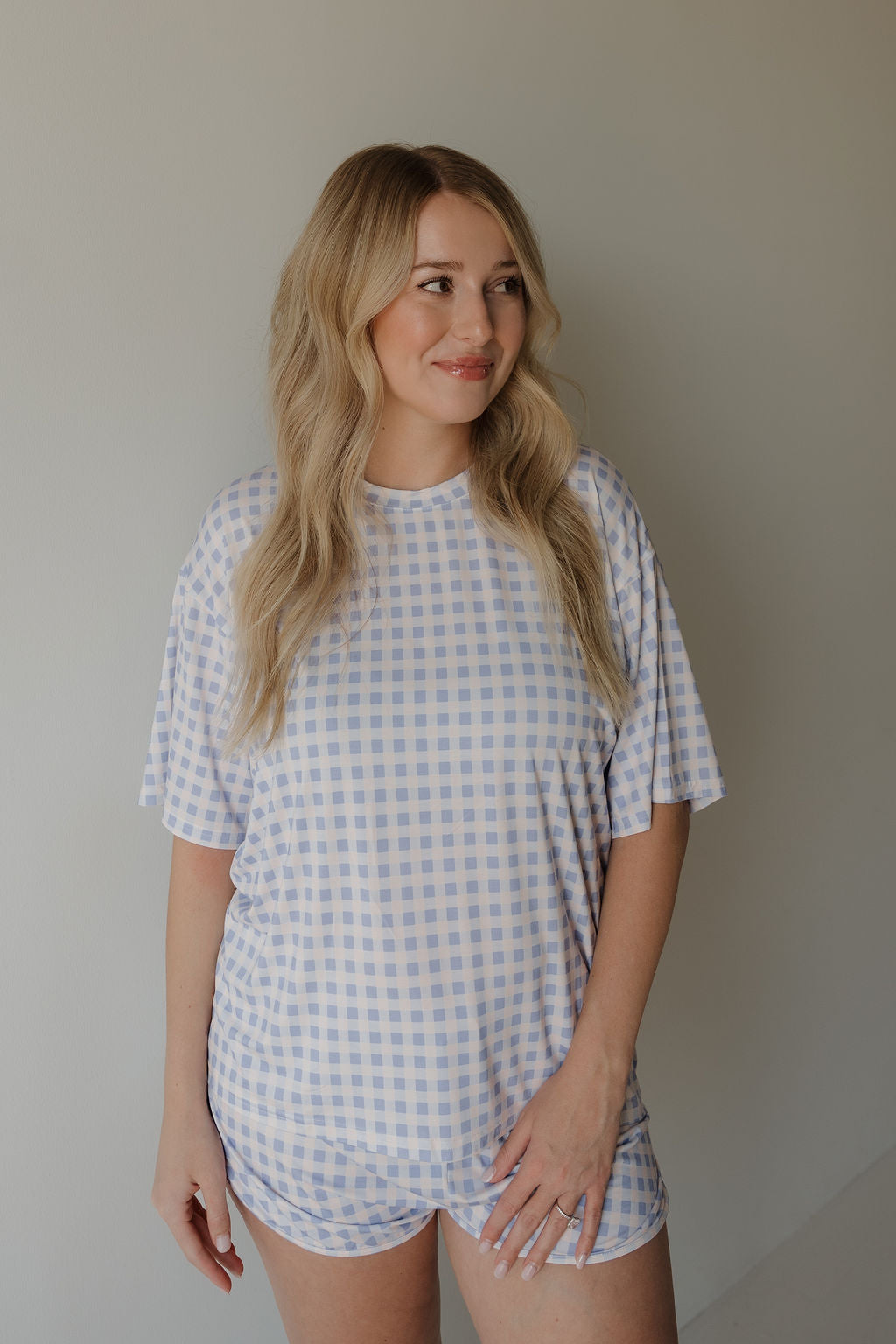 A woman with long blonde hair stands against a plain light background, smiling slightly to the side while wearing the forever french baby Women’s Bamboo Short Set | Marilyn—a matching light purple and white checkered short-sleeve top and shorts made from breathable bamboo fabric.
