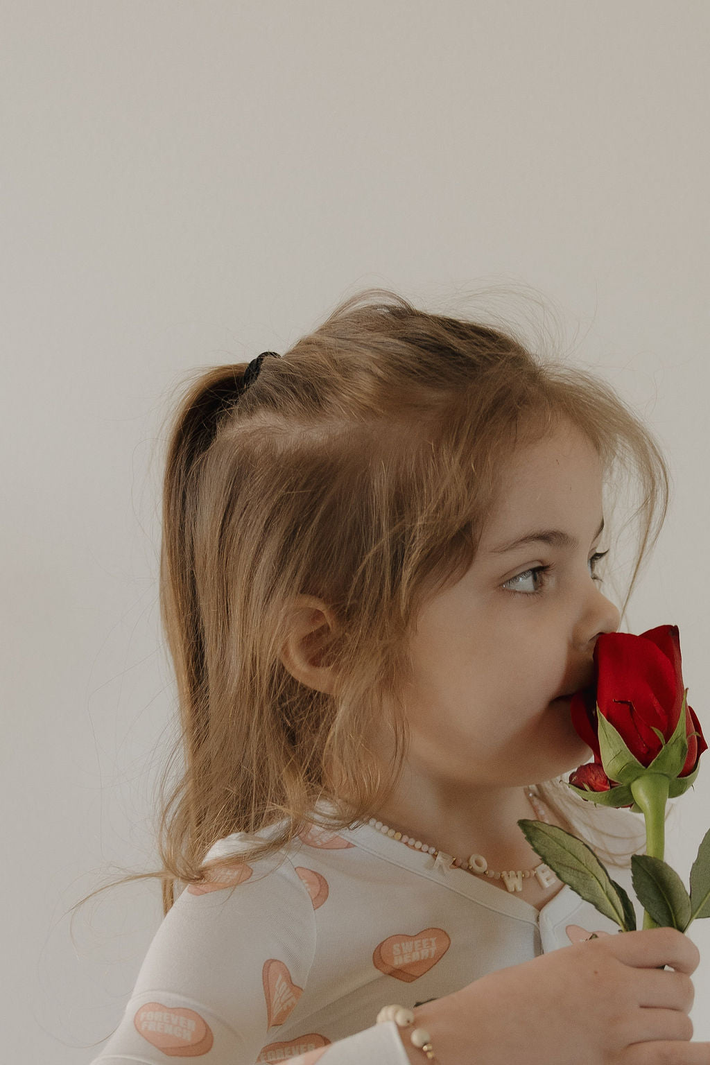 A young girl with light brown hair in a ponytail smells a red rose, looking to the side. She wears forever french baby's Bamboo Two Piece Pajamas in Love Notes - Grey, and a beaded necklace, standing against a plain light background.