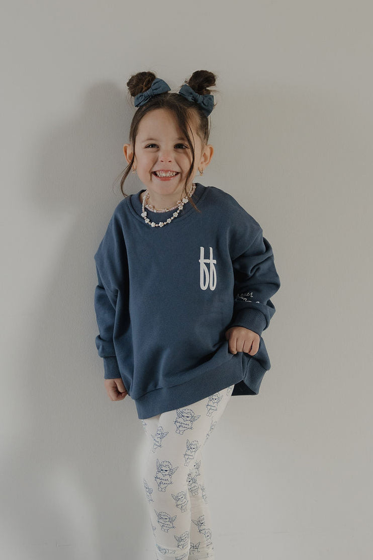 A smiling young girl with brown hair in two buns wears the forever french baby Child Sweatshirt | ff Blue, white leggings with a blue floral print, a beaded necklace, and a blue bow headband, standing by a plain light background.