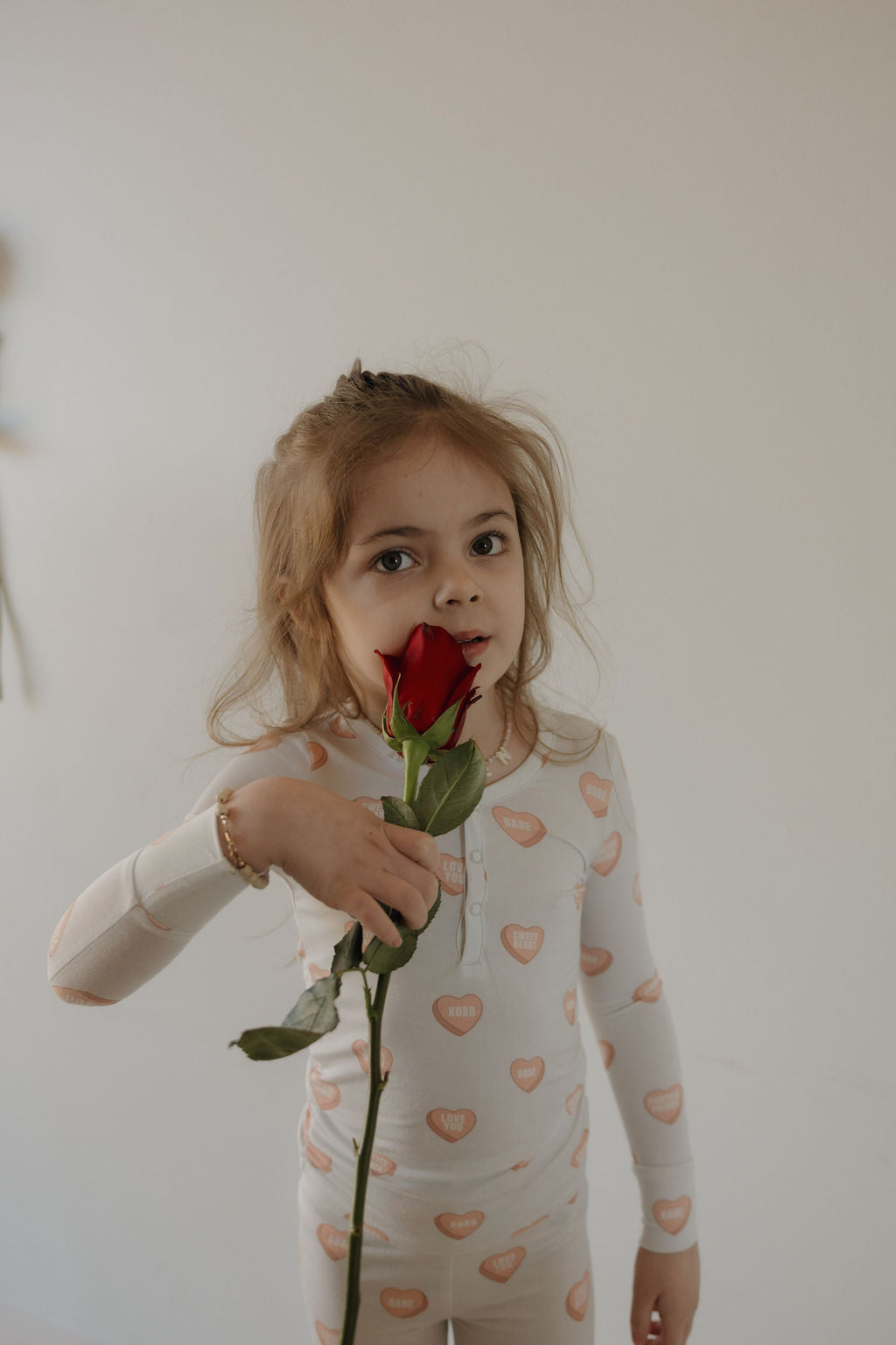 A young girl with long light brown hair holds a red rose close to her face, wearing forever french baby Bamboo Two Piece Pajamas | Love Notes - Grey, featuring breathable kids fabric and pink heart patterns against a plain light-colored wall.