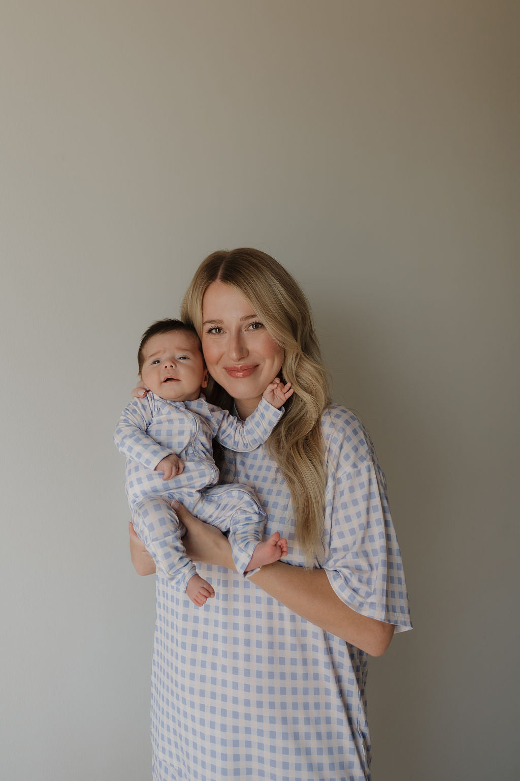 A woman with long blonde hair holds a baby, and both are dressed in matching blue and white checkered hypo-allergenic loungewear. The woman is wearing the Women’s Bamboo Short Set | Marilyn by forever french baby, as they stand against a plain light-colored background.