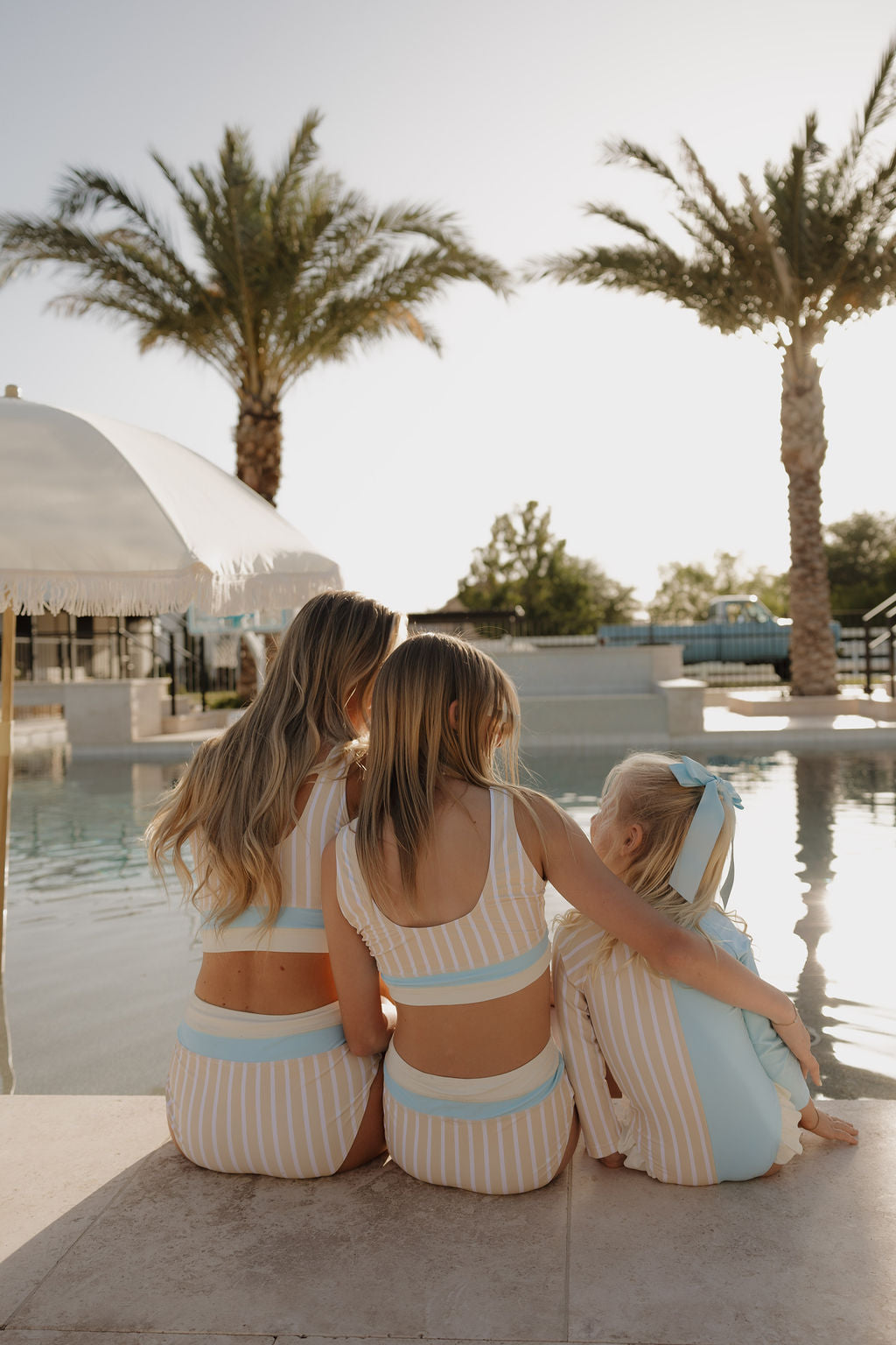 Three girls in matching lolo webb 2 Girl Long Sleeve Swimsuit | Surf & Sand sit poolside with their backs to the camera, enjoying the sun. In the background are palm trees and a white umbrella. The smallest girl wears a bow in her hair.