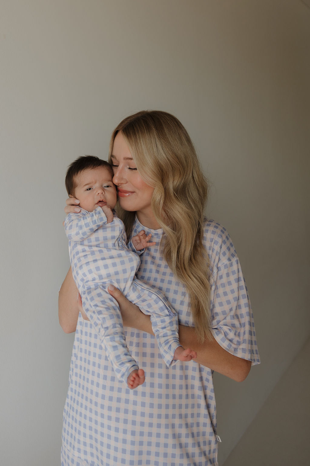 A woman with long blonde hair holds a baby against a neutral background. Both are wearing matching forever french baby Bamboo Zip Pajamas | Marilyn in pastel gingham, made from breathable fabric. The woman smiles gently with her eyes closed as the baby looks toward the camera.