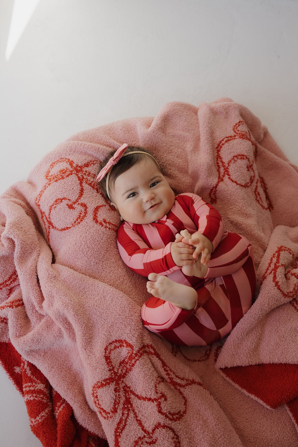 A baby in a red and pink striped outfit with a pink bow headband smiles while lying on the Minty Cherries Plush Blanket | Iconic Cherry, featuring soft fabric and playful cherry-themed patterns.